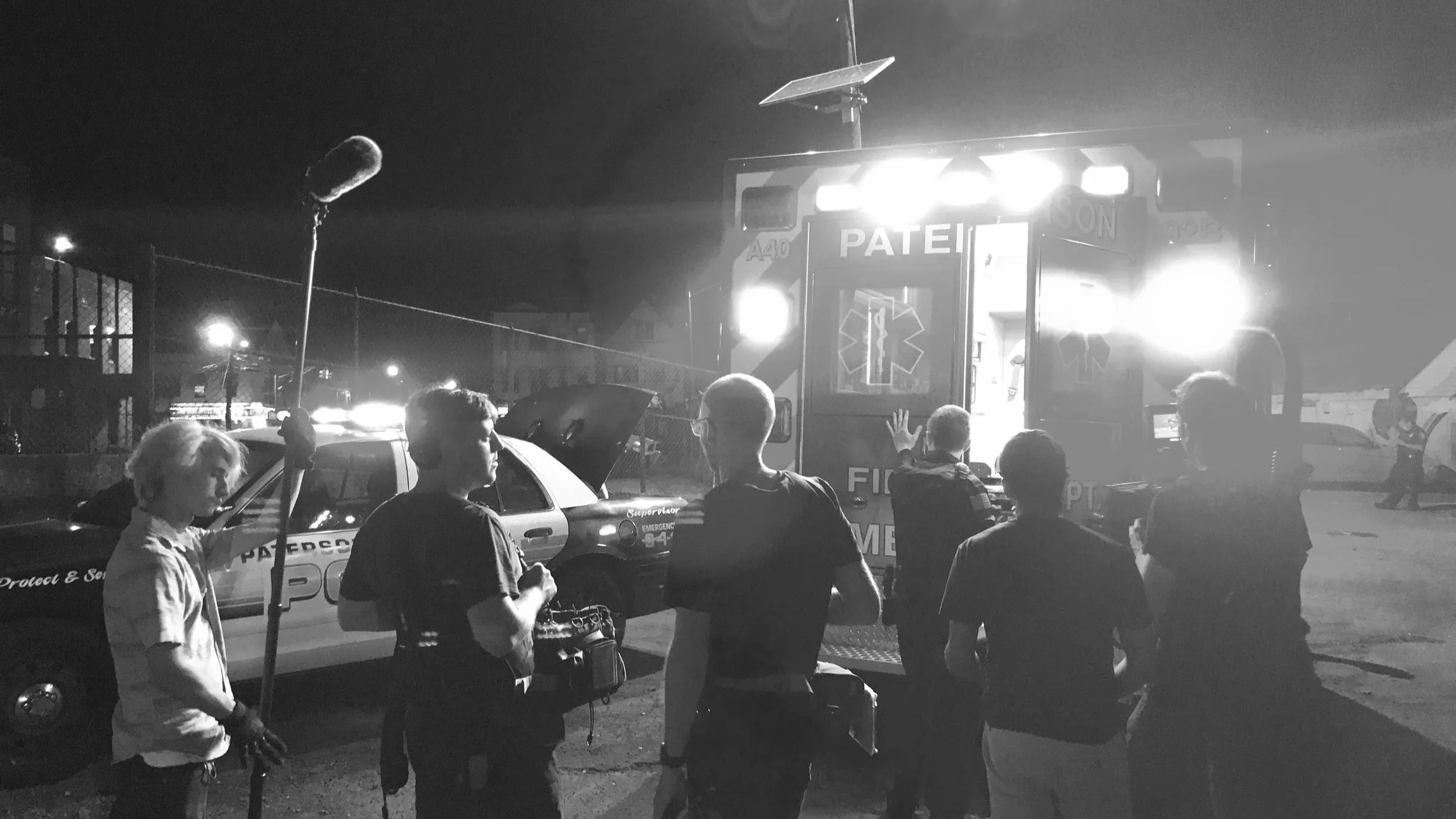 Nighttime scene of emergency responders, including police officers and paramedics, gathered near an ambulance with lights on, parked next to a police car with open compartments. Several people are standing and talking, with a microphone visible in th