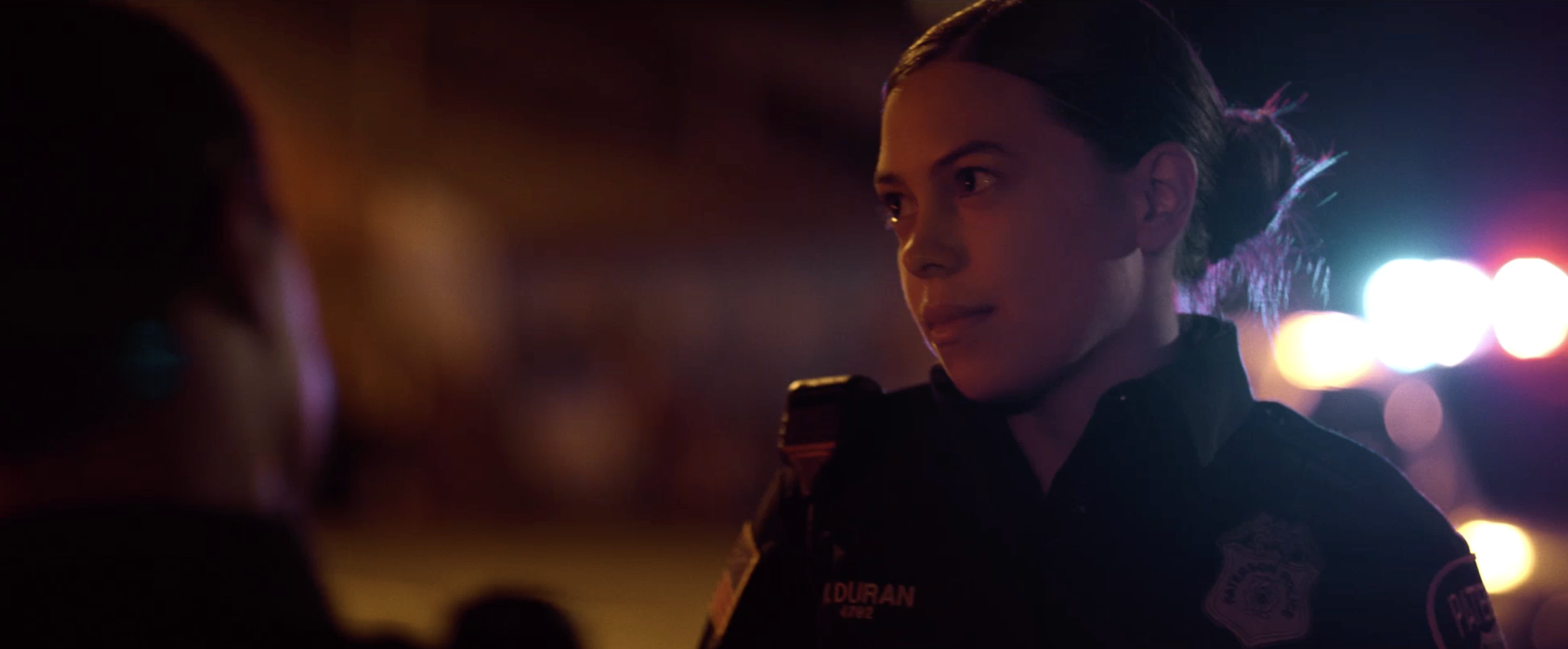 A female police officer with a serious expression talking to someone at night, with city lights and bokeh in the background.