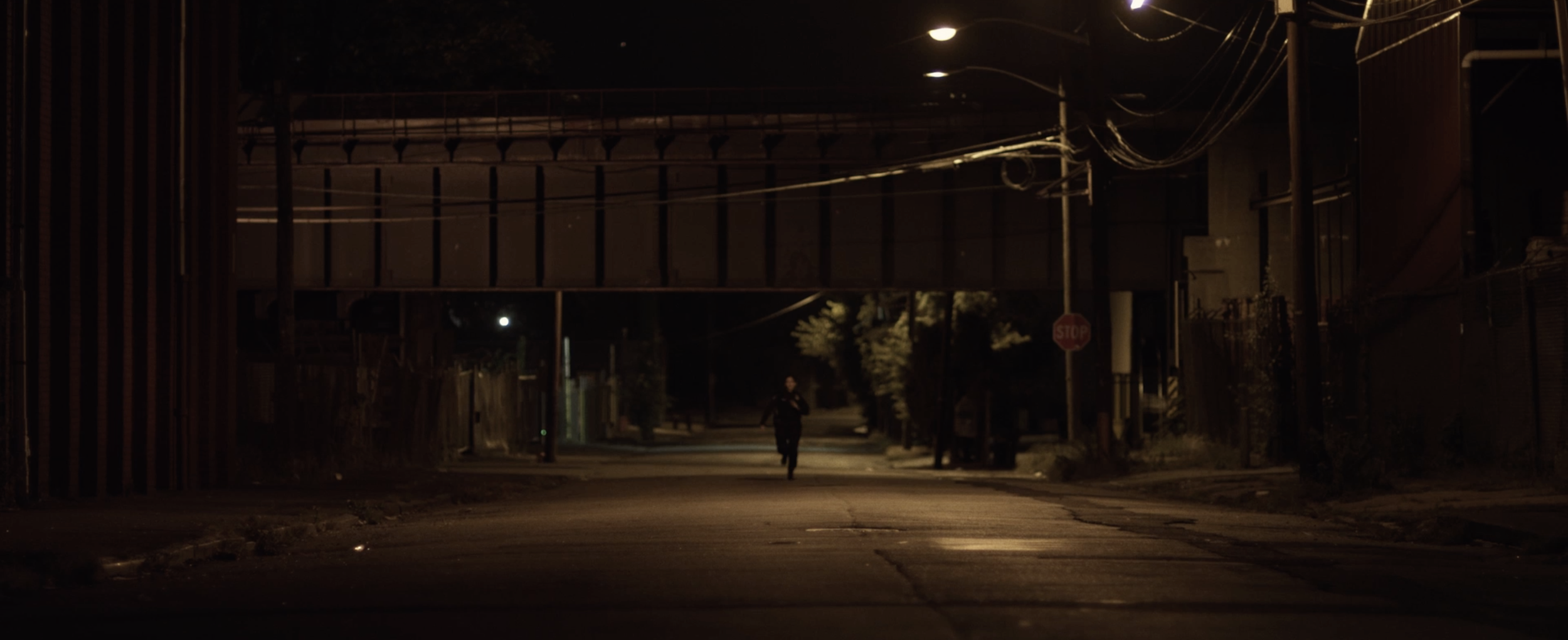 A person jogging alone on a dimly lit street at night, with an overpass in the background and a stop sign visible on the right side.