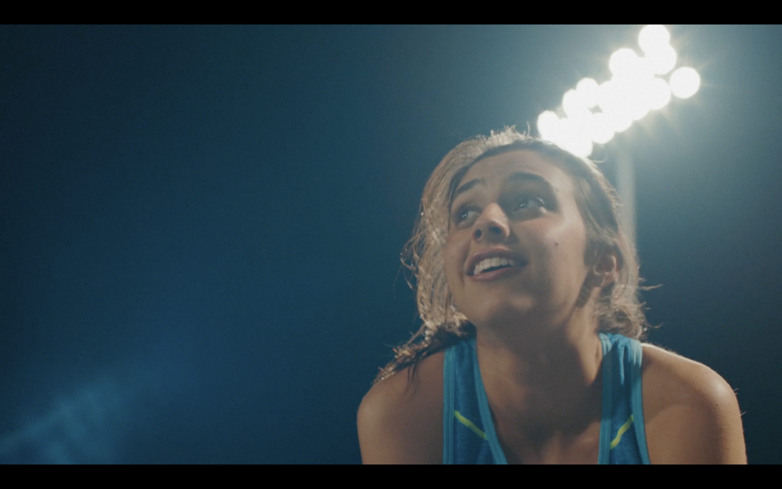 A woman with short blonde hair, wearing a blue athletic tank top, smiling and looking upward, with bright lights in the background.