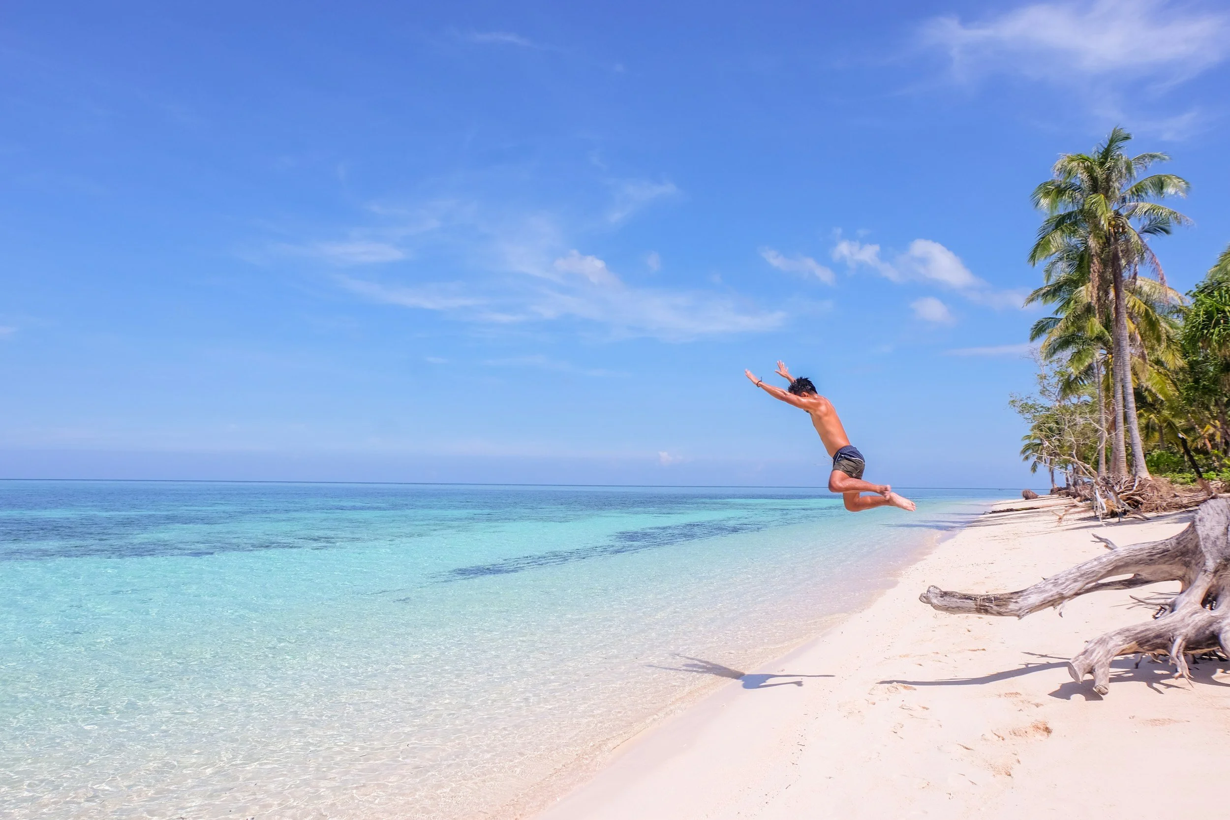 A person jumping on a tropical beach with clear blue water, white sand, palm trees, and a partly cloudy sky.