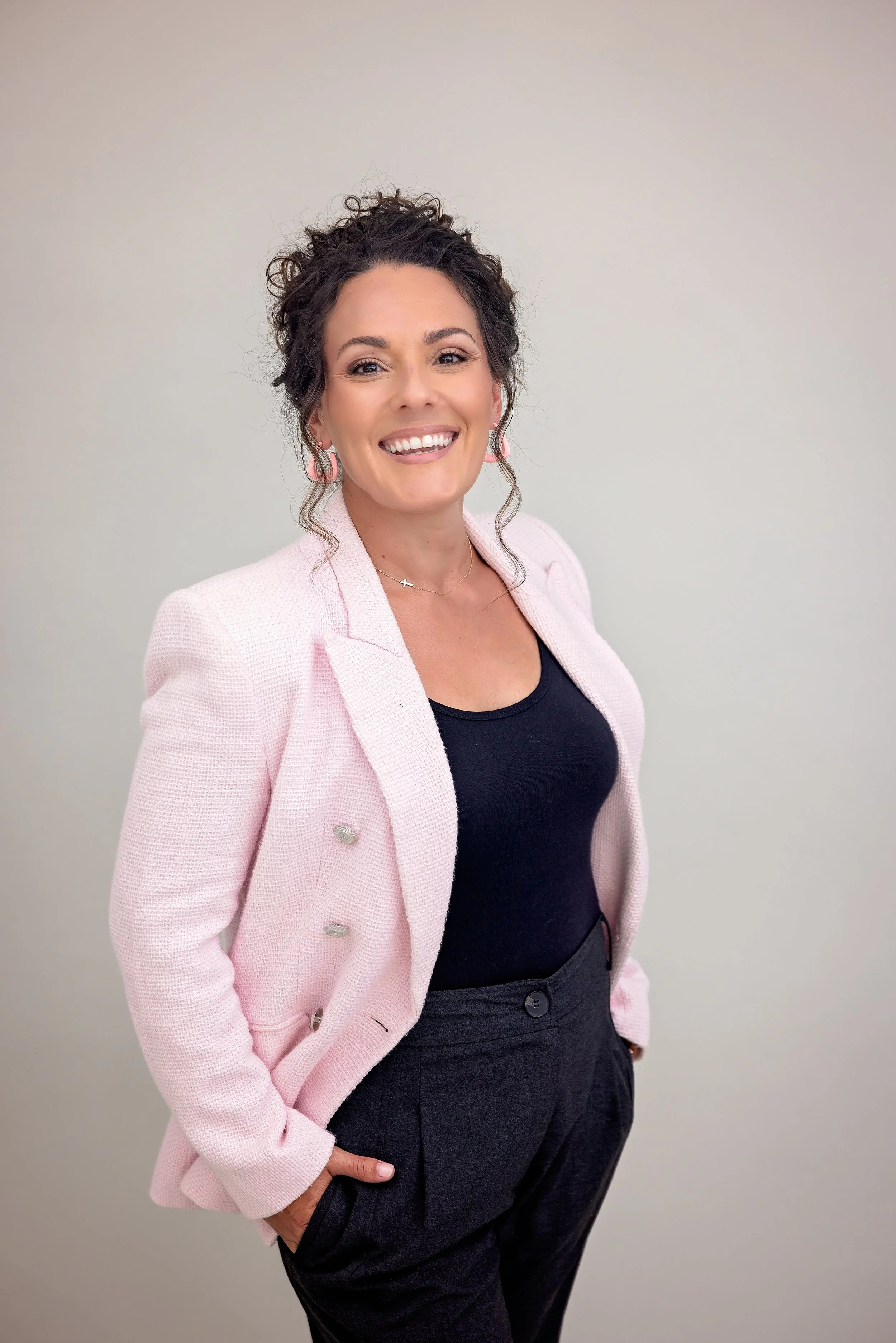 A woman with curly dark hair is smiling and looking at the camera. She is wearing a light pink blazer, a black top, and dark pants. She is standing against a plain light-colored background.