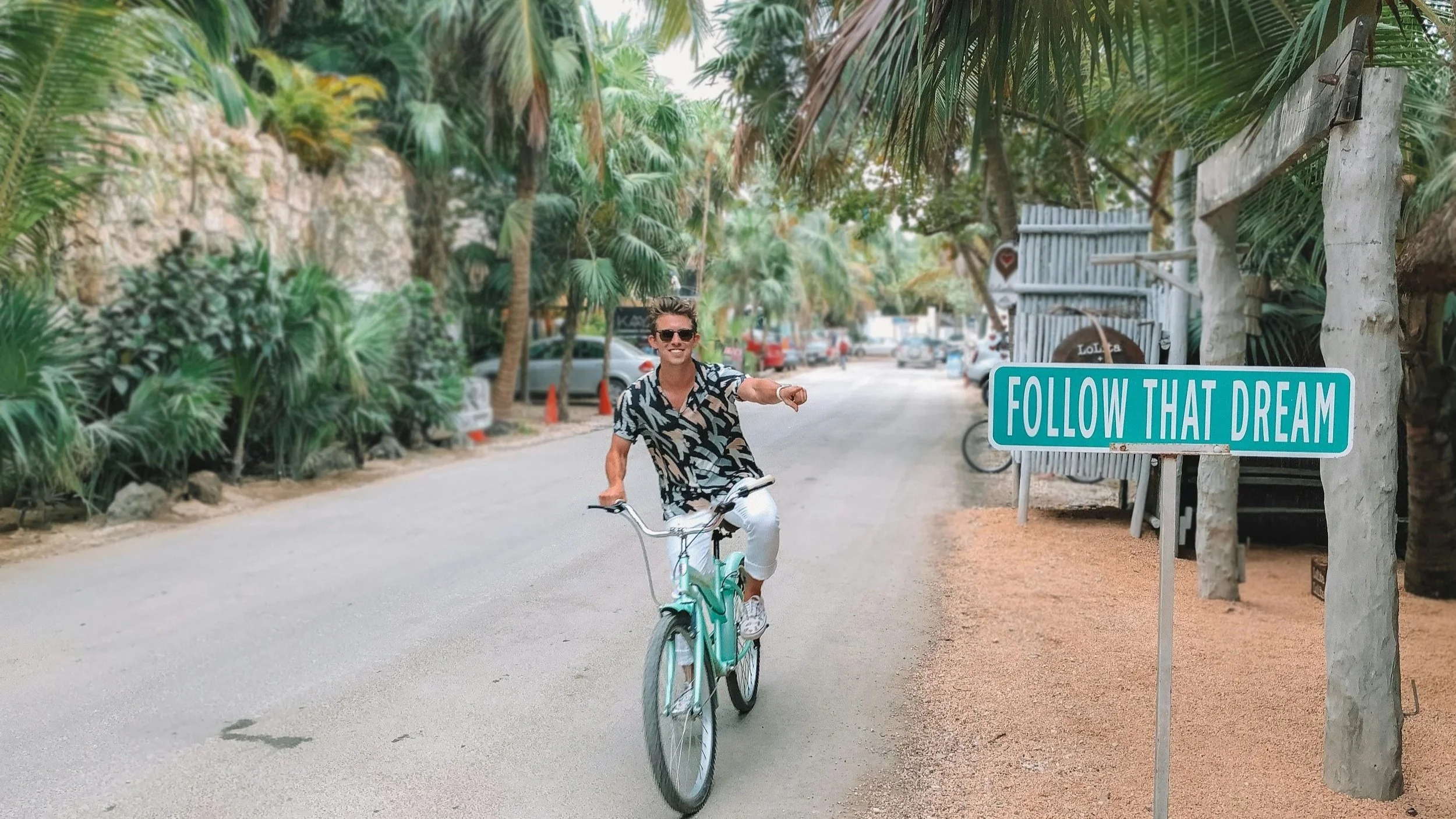 A man riding a bicycle on a tropical street with palm trees, wearing sunglasses and a patterned black and white shirt, smiling. A sign reads 'FOLLOW THAT DREAM.'