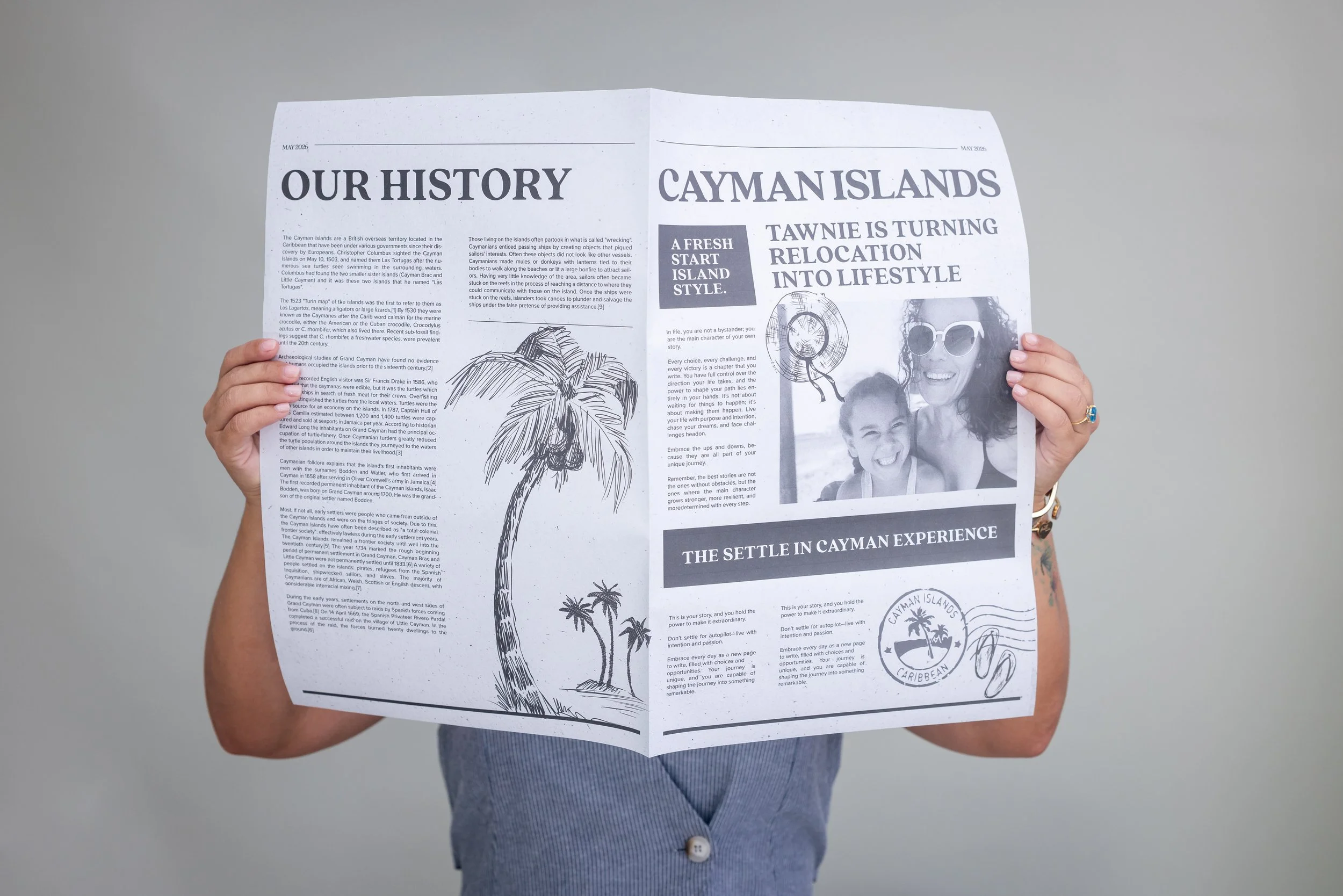 A person holding up a black-and-white newspaper titled 'Our History Cayman Islands' in front of a gray background, with part of their arm and a portion of their clothing visible.