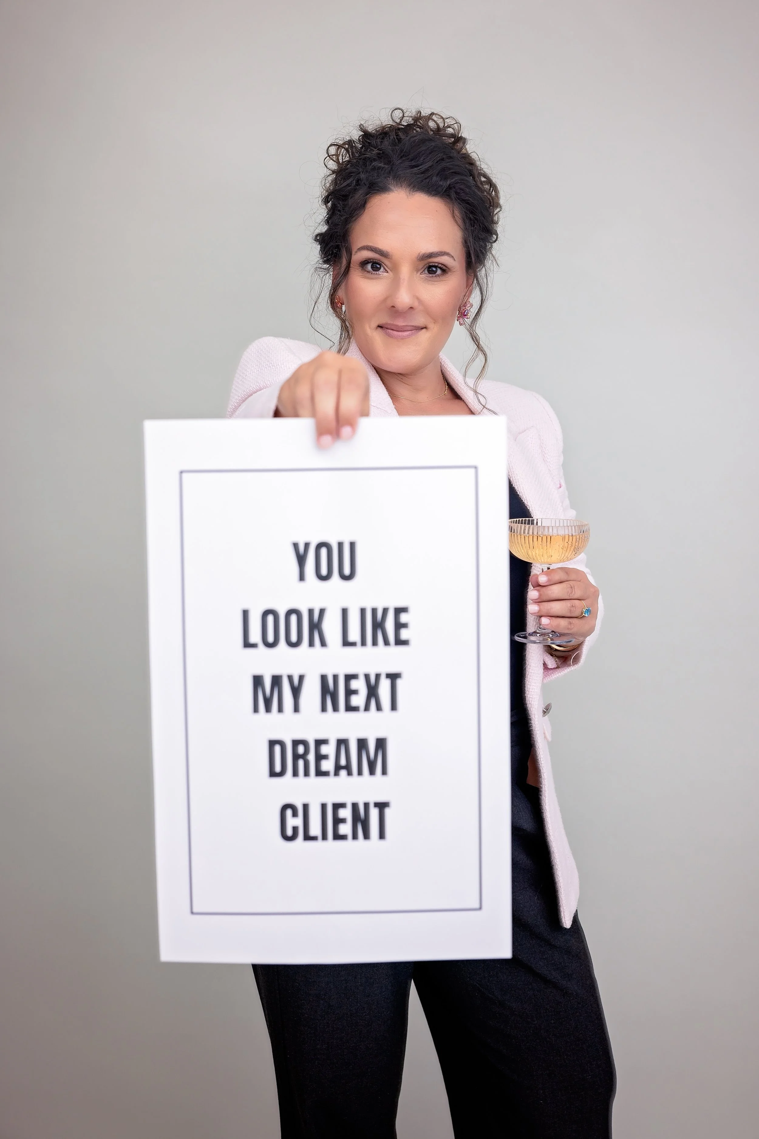 A woman with curly hair and earrings holding a glass of wine in one hand and a sign with the message 'You look like my next dream client' in the other.