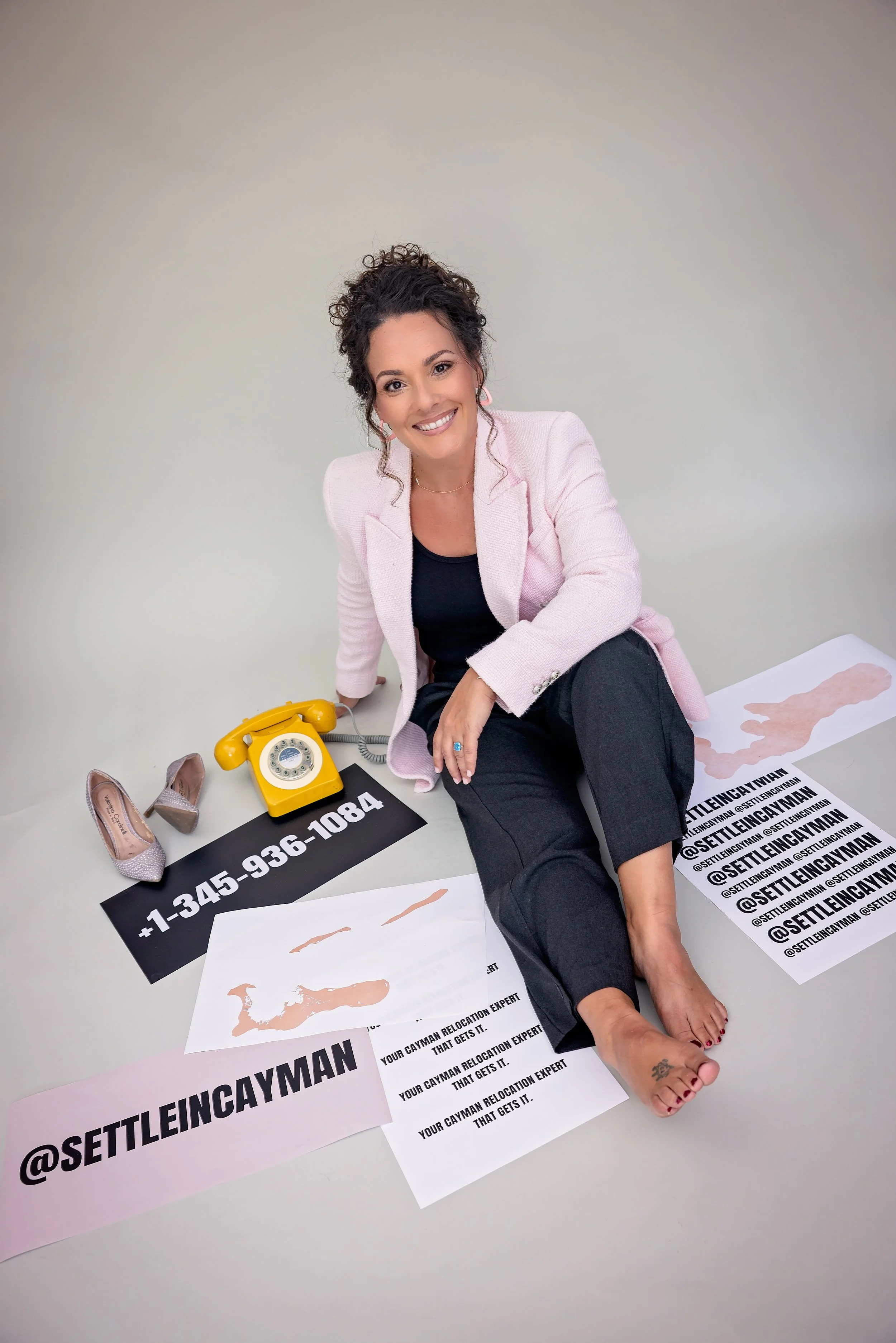 A smiling woman with dark curly hair, wearing a pink blazer and black pants, sitting on the floor surrounded by papers with the handle @SETTLEINCAYMAN, a yellow rotary phone, and a pair of heels. The papers display a map of Cayman and phone numbers.