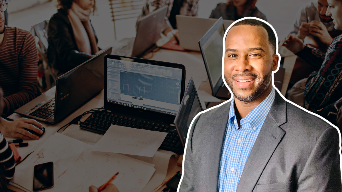 A man in a suit smiling in front of a background of a business meeting with people working on laptops.