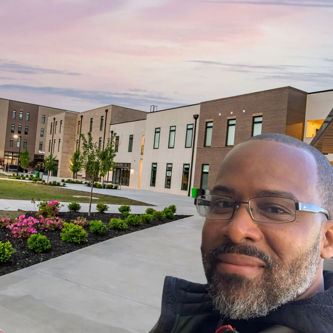 A man with glasses and a beard taking a selfie outdoors with modern buildings, trees, and pink flowers in the background during dusk.
