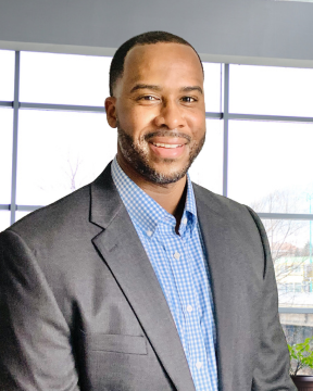A professional man in a gray blazer and blue checkered shirt standing indoors with large windows in the background.