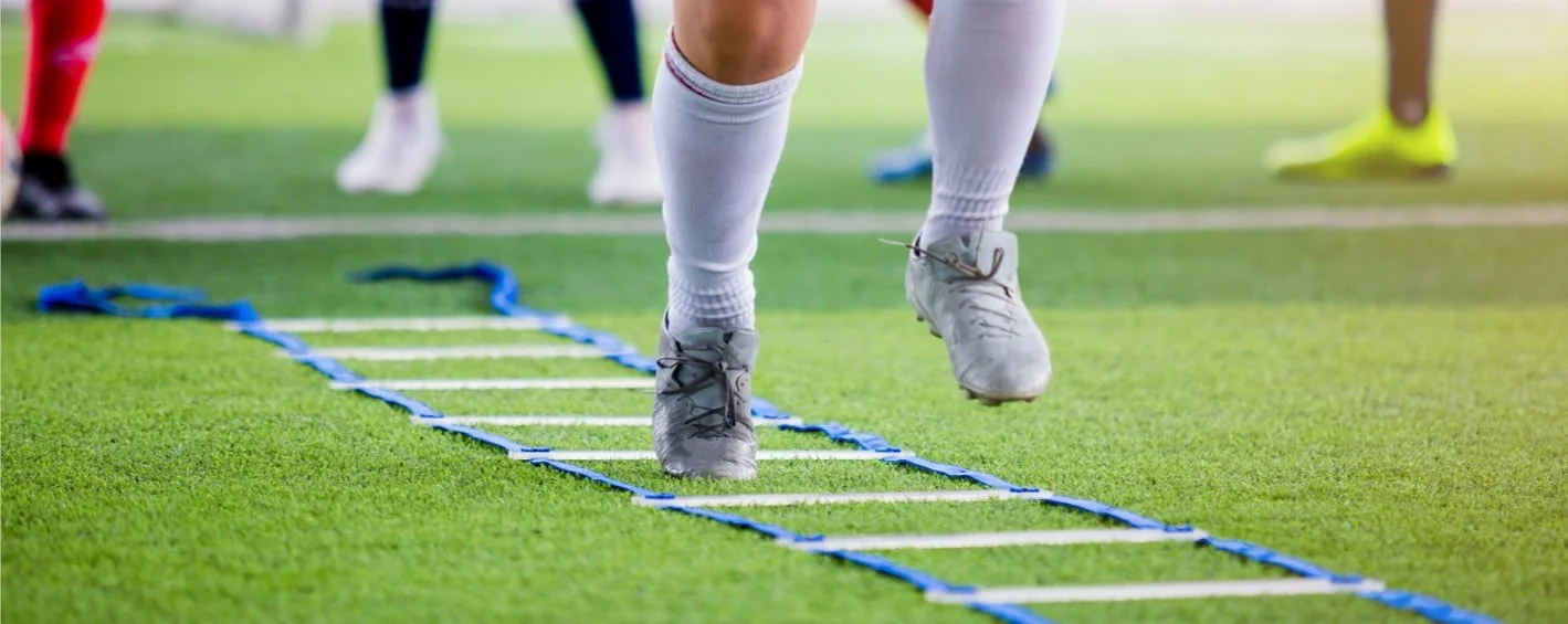 Close-up of a person in athletic shoes and white socks performing agility drills on a grassy field with a blue agility ladder.