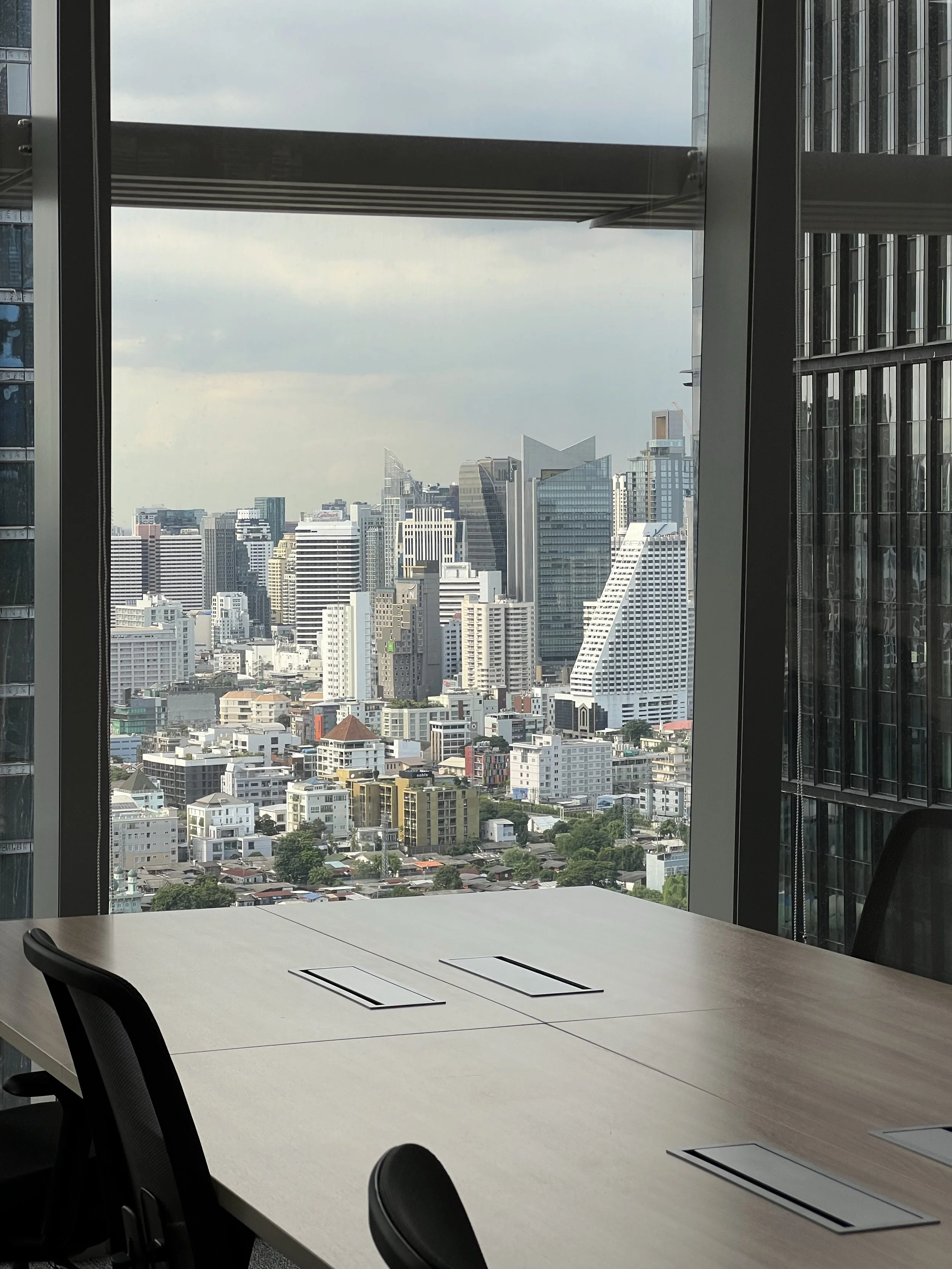 Office conference room with a large window overlooking a city skyline of tall buildings under a cloudy sky.