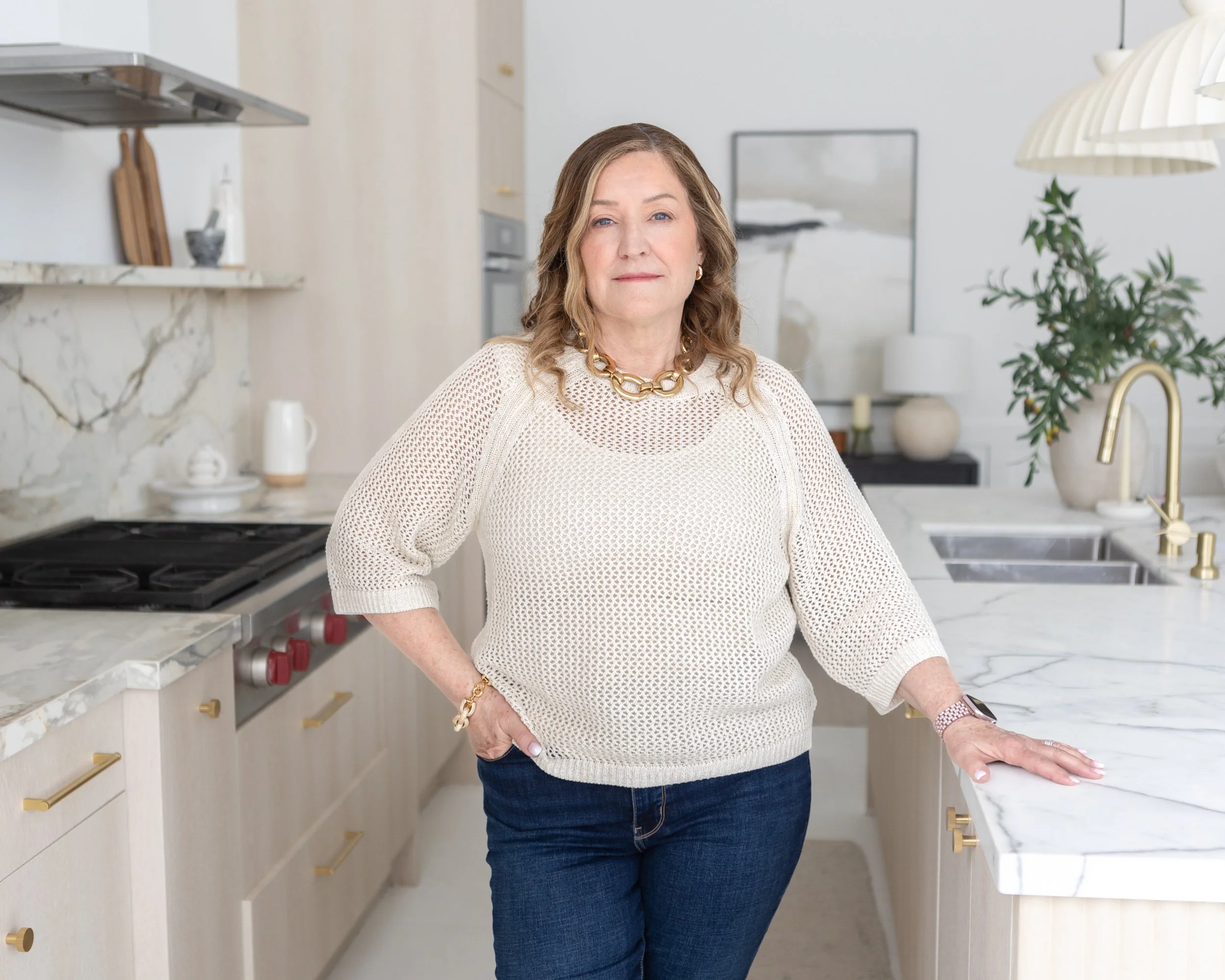 A woman with wavy light brown hair wearing a white knitted sweater, gold chain necklace, and watch, standing in a modern kitchen with marble countertops, white cabinetry, and a potted plant.