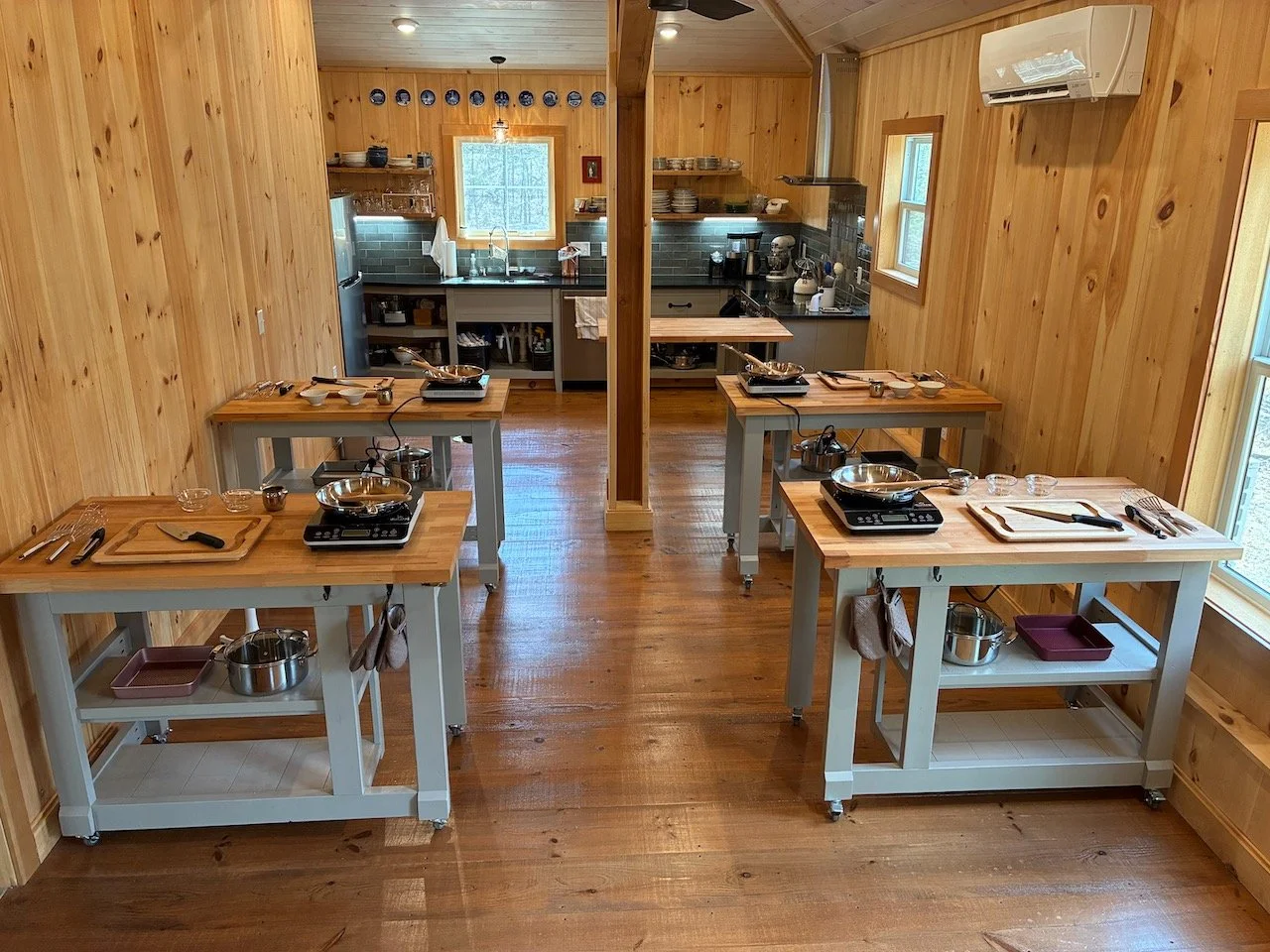 A farm style kitchen classroom featuring four cooking stations with induction burners, pans, and utensils.