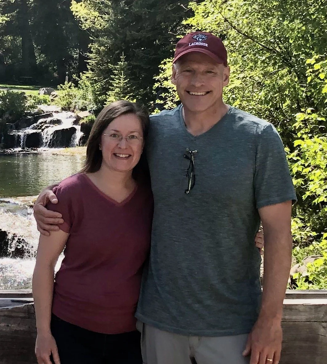 A smiling woman and man standing outdoors surrounded by trees and greenery.