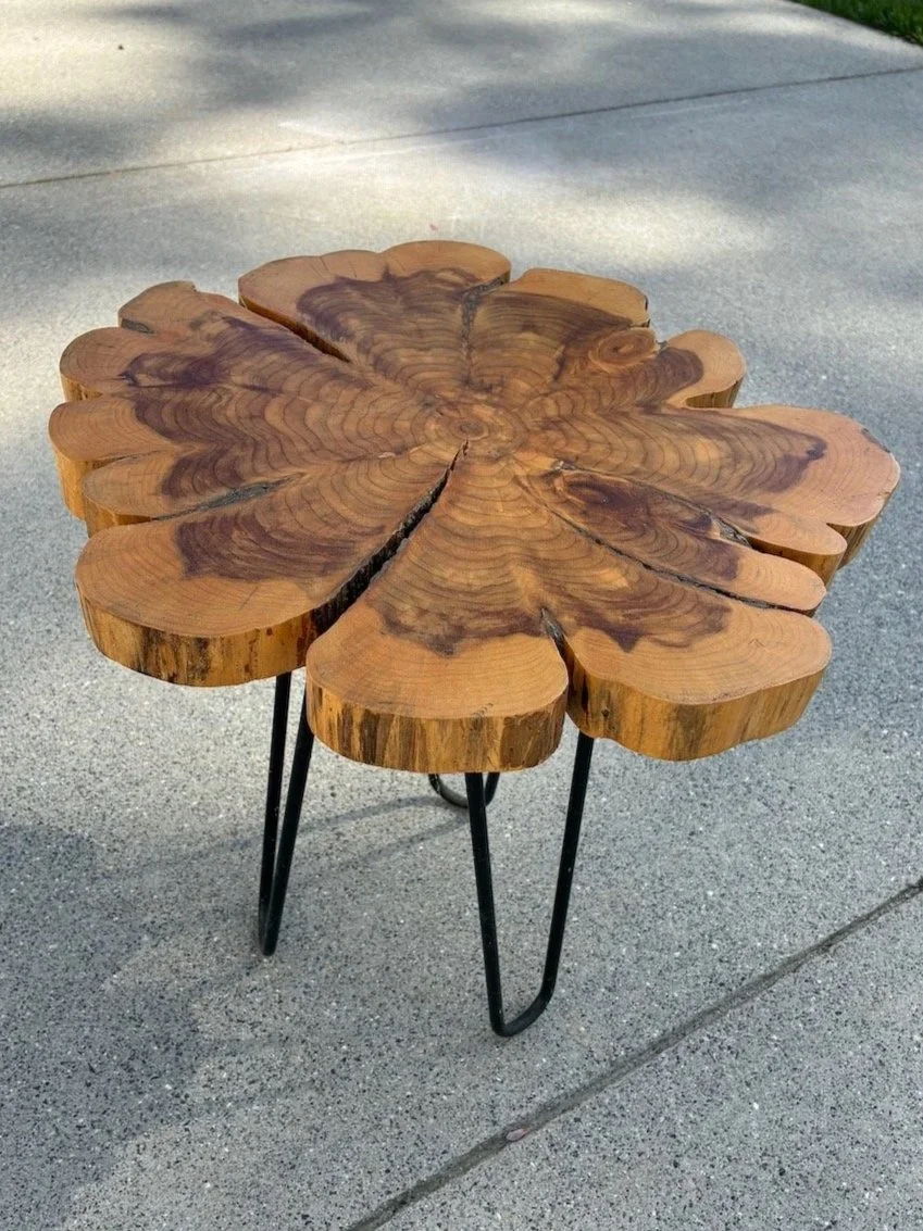 A rustic side table made from a cross-section of a tree trunk with a flower petal shape and black metal hairpin legs.