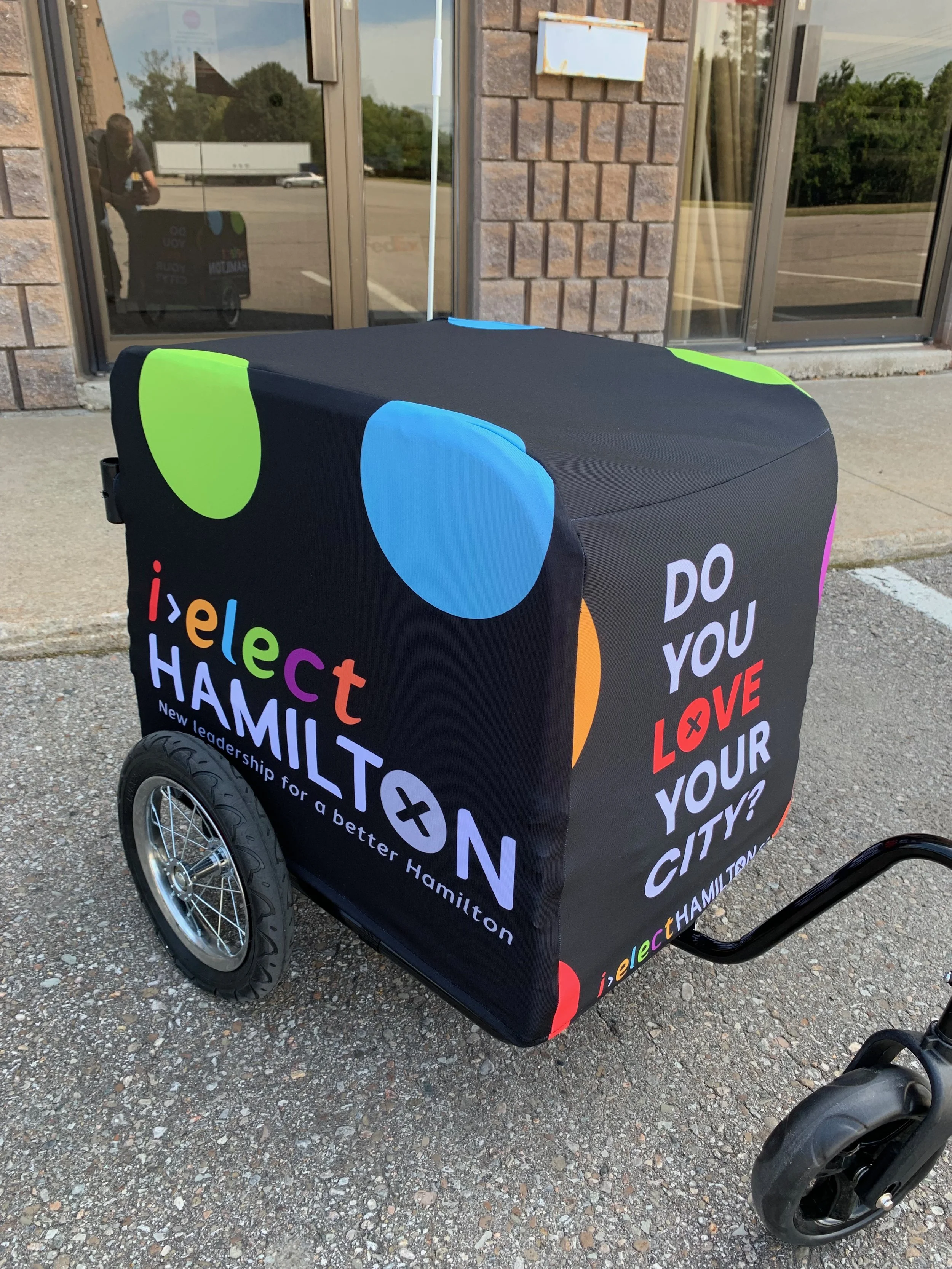 A stretchy black cover that reads "i>elect Hamilton" on the side and "Do you love your city?" on the front covering a wheeled cart