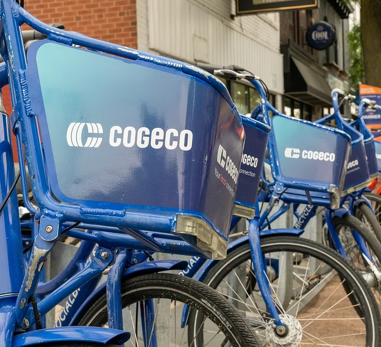 Blue bikes parked at a storage rack that have a vinyl display installed that has the Cogeco logo on the basket