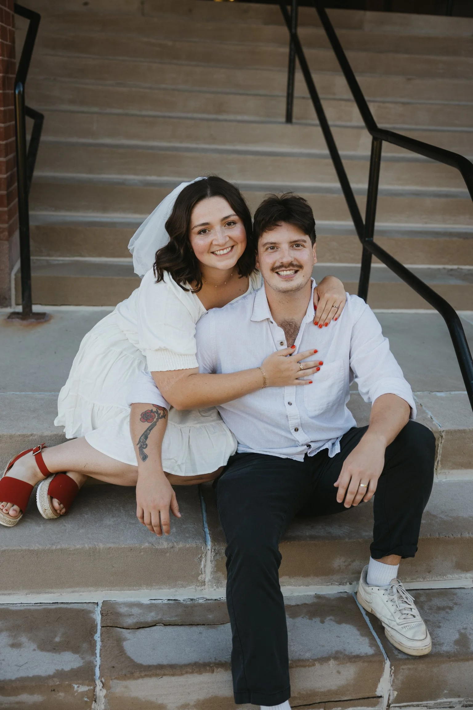 A young woman with dark wavy hair, wearing a white dress and red shoes, sits on steps with a man in a white shirt and black pants. They are smiling, and she has her arm around his shoulder while he has one hand on his knee. The woman has a tattoo on her left forearm and is wearing red nail polish. The scene appears to be outdoors, in front of a building with stairs and black metal railings.