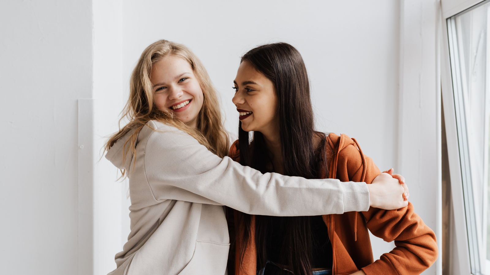 Two Women hugging and laughing. They are inside a house with a white wall.