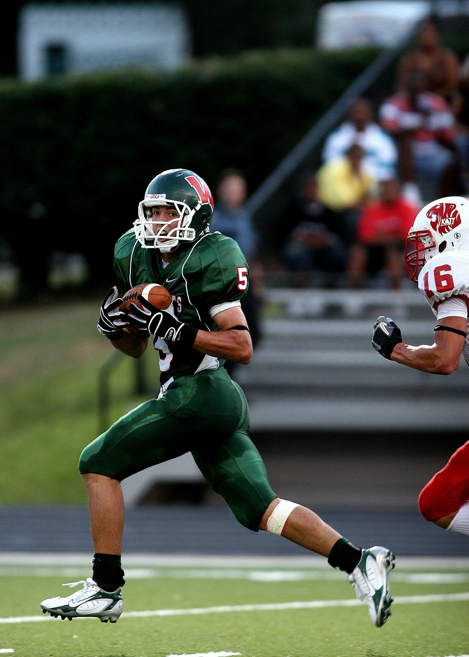 A football player in a green uniform with the number 5 on his jersey running with the ball during a game, with an opposing player in a white and red uniform chasing him.