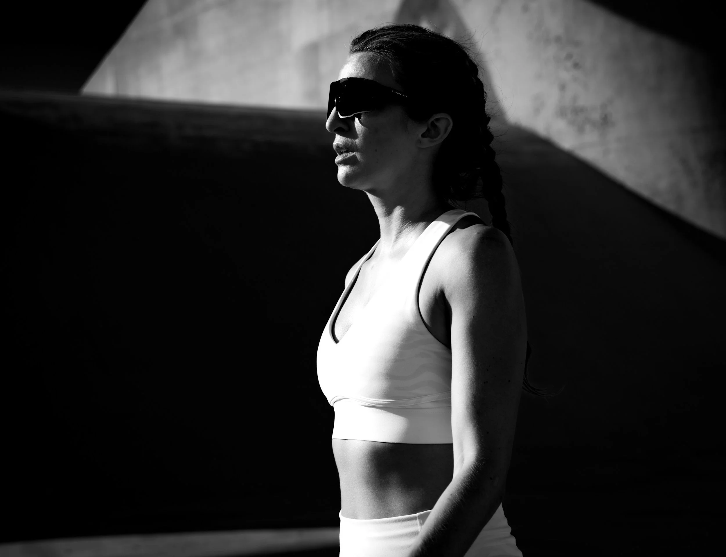 A woman in athletic wear with sunglasses standing outdoors in side profile, with high-contrast lighting creating shadows.