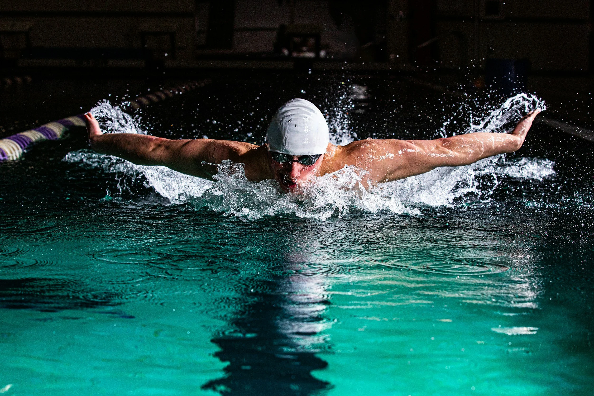 Swimmer performing a butterfly stroke in an indoor swimming pool, wearing a cap and goggles.