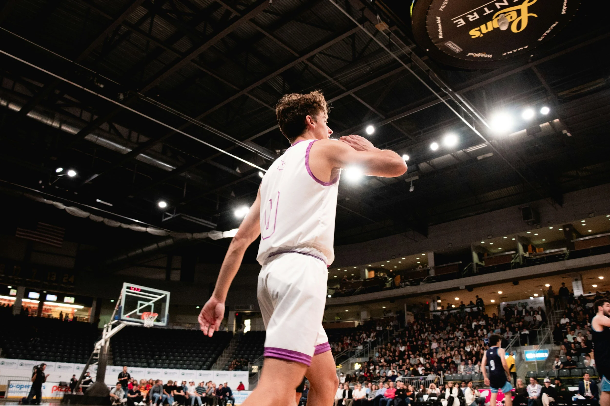A basketball player in a white uniform with purple accents is on the court during a game, with crowd and other players visible in the background.