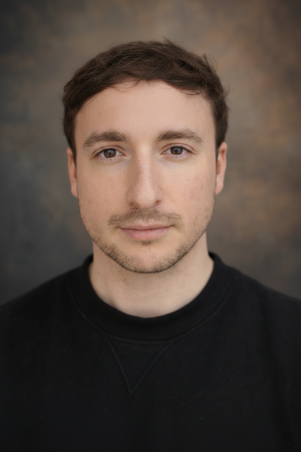 A young man with brown hair, light facial hair, and fair skin, wearing a black shirt, looking directly at the camera with a neutral expression, against a blurred background.