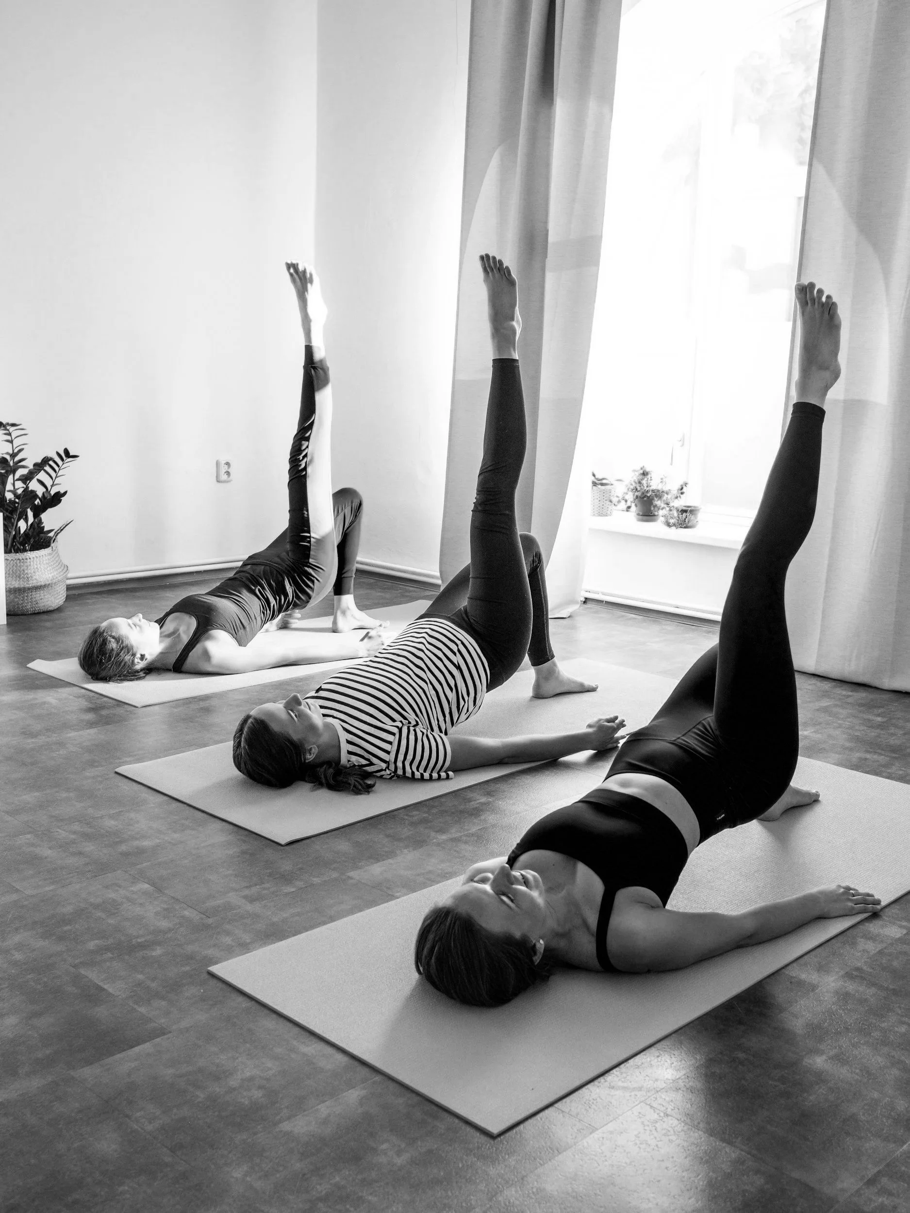 Four women doing yoga poses on mats in a well-lit room with wooden floors, a window with curtains, and potted plants.