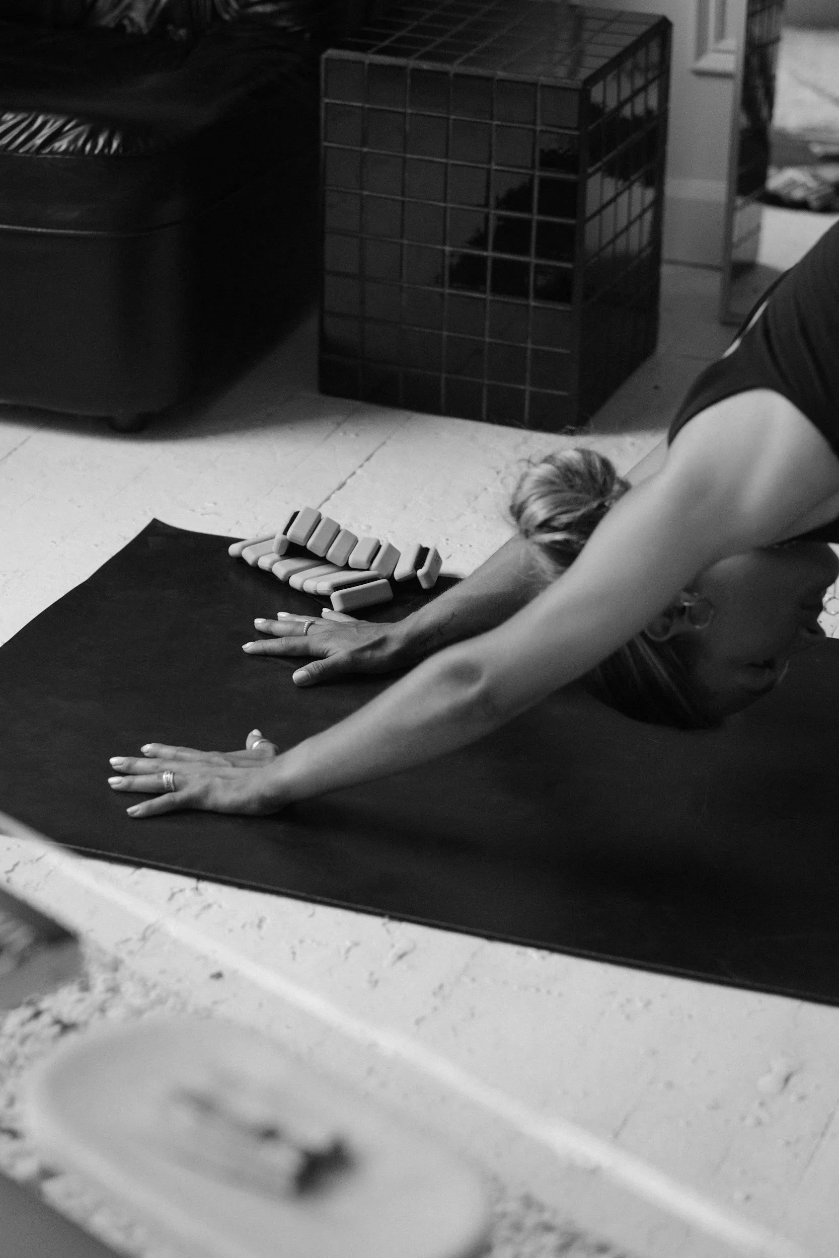 A person practicing yoga or stretching on a black mat, with domino tiles arranged in a line in front of them. They are in a room with a black storage bin and a cage-like item in the background.