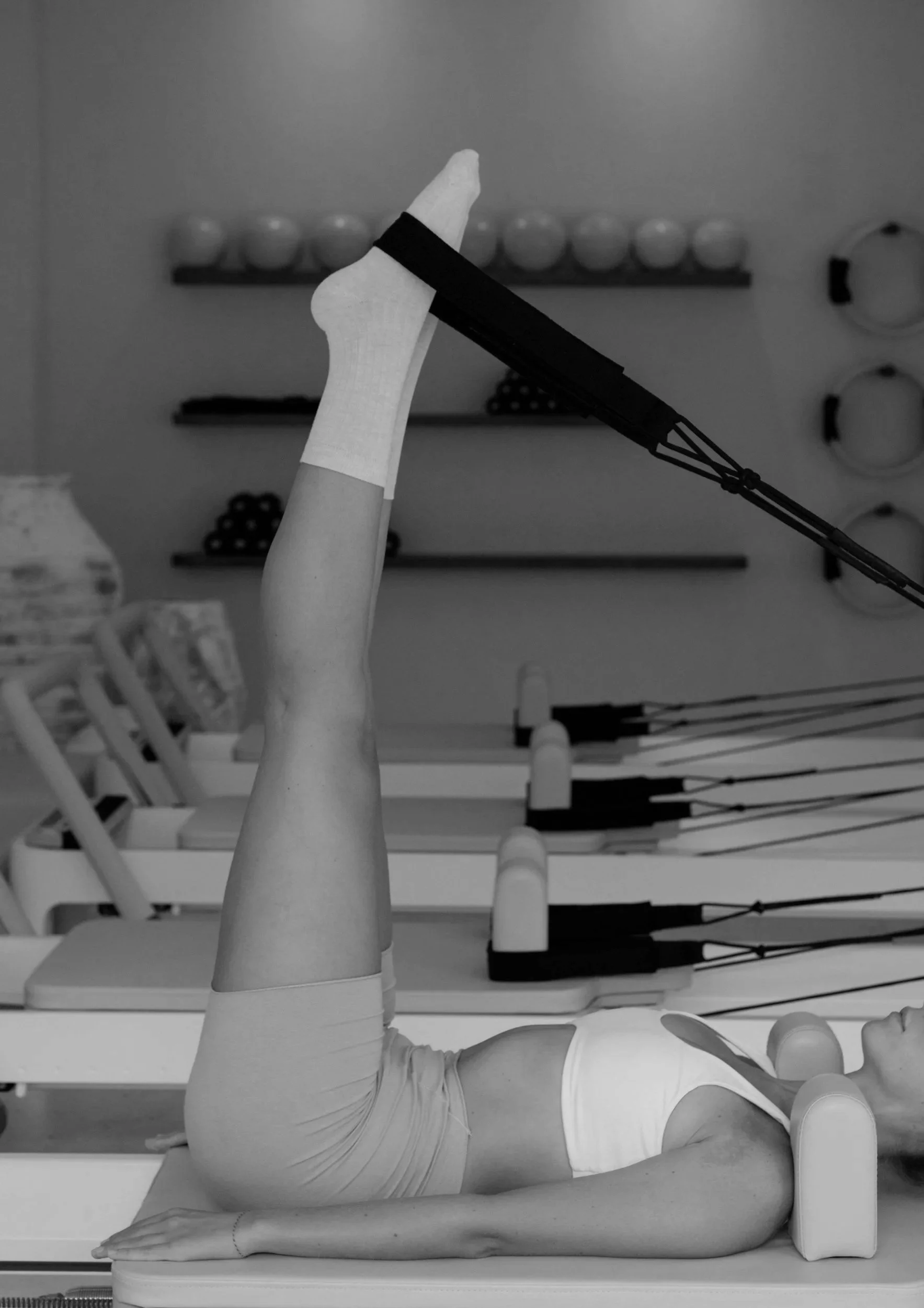 A person lying on a flat surface, head turned to the side, with one leg raised and bent, holding a yoga or stretching pose against a plain background with shelves and various objects.