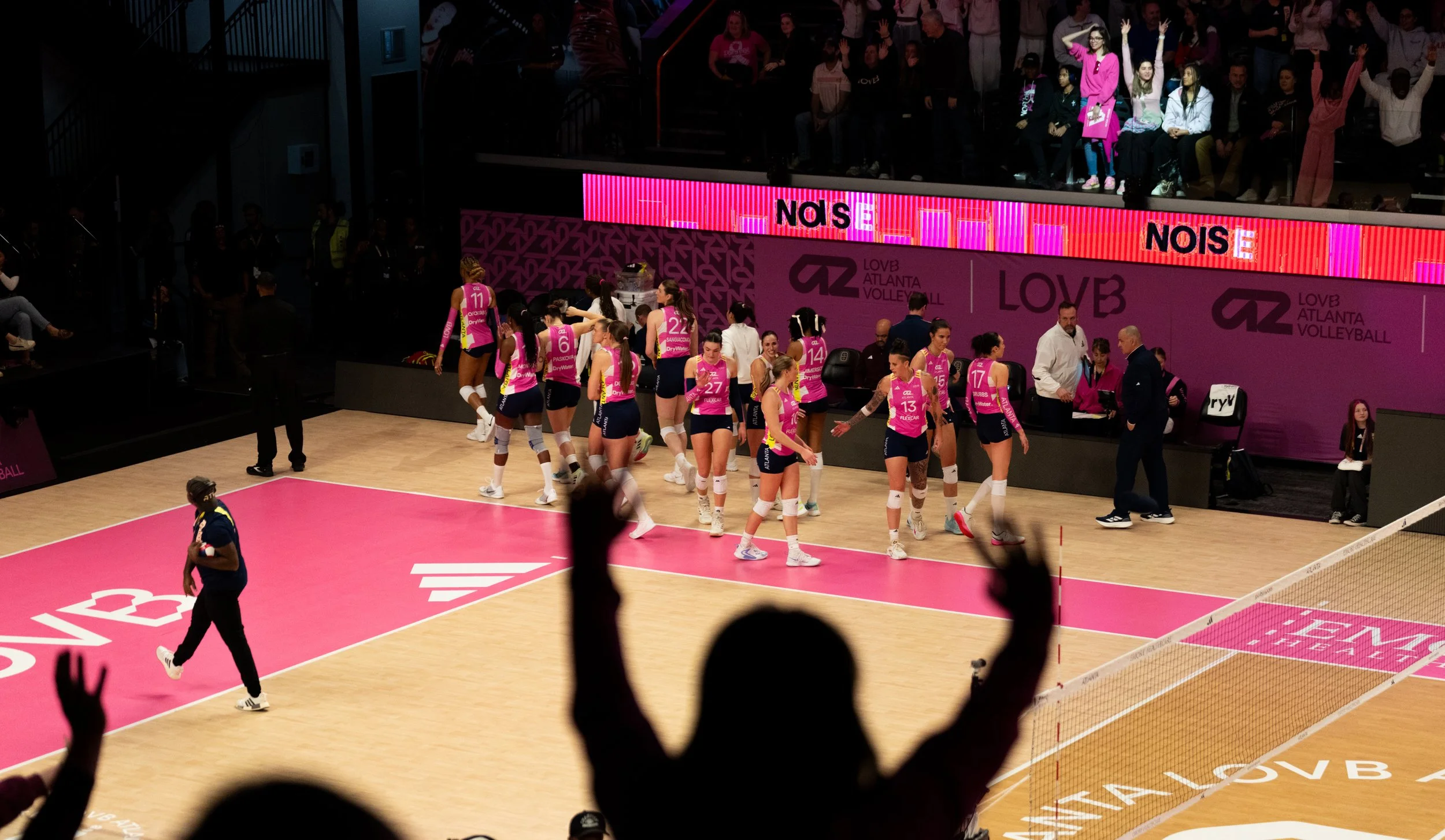 A women's volleyball team, wearing pink and blue uniforms, celebrates on the court after a match at a volleyball arena. Spectators are visible in the background, and members of the team are shaking hands while some raise their hands in celebration.