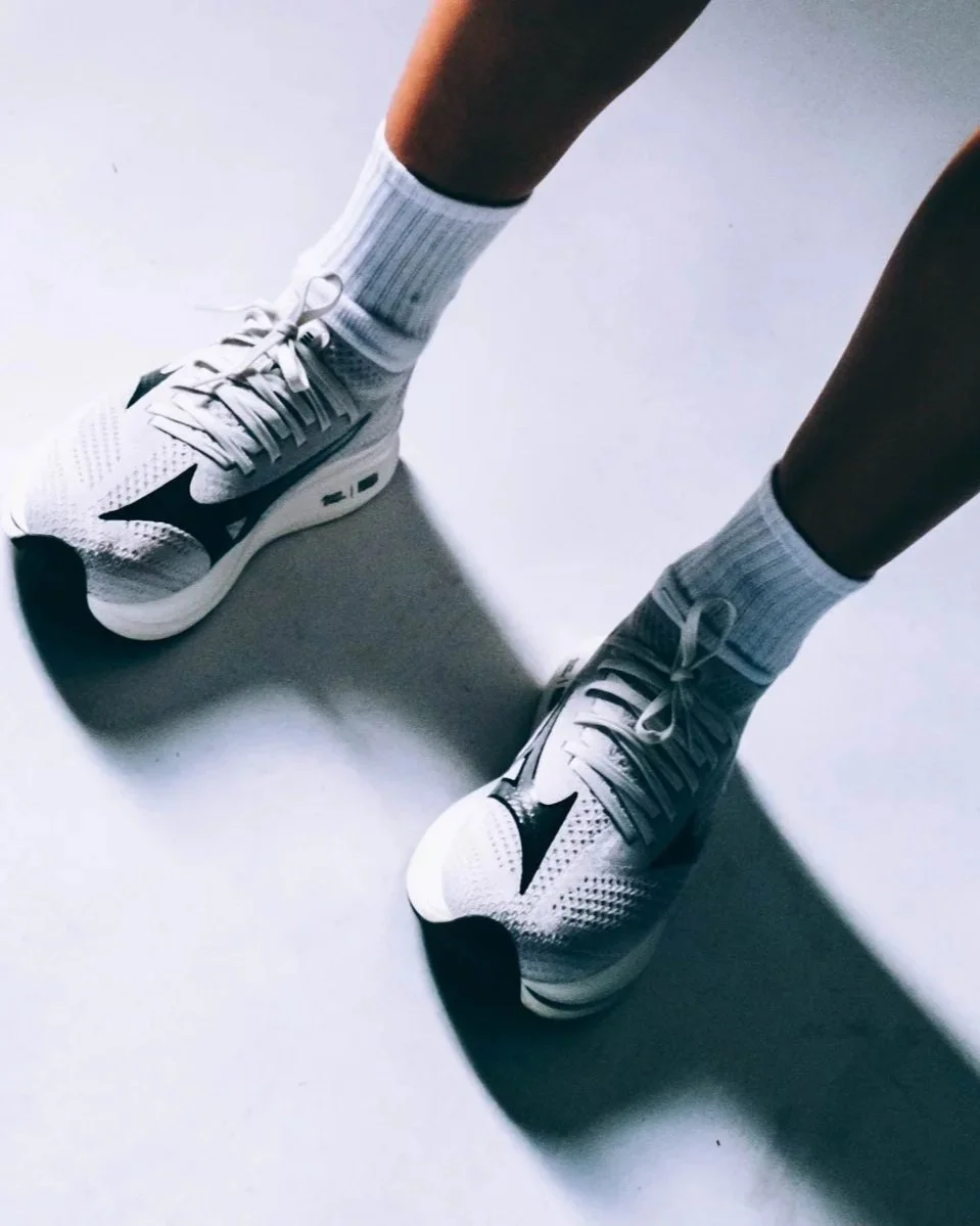 Person wearing white athletic shoes with black swoosh logos and white socks, standing on a white surface.