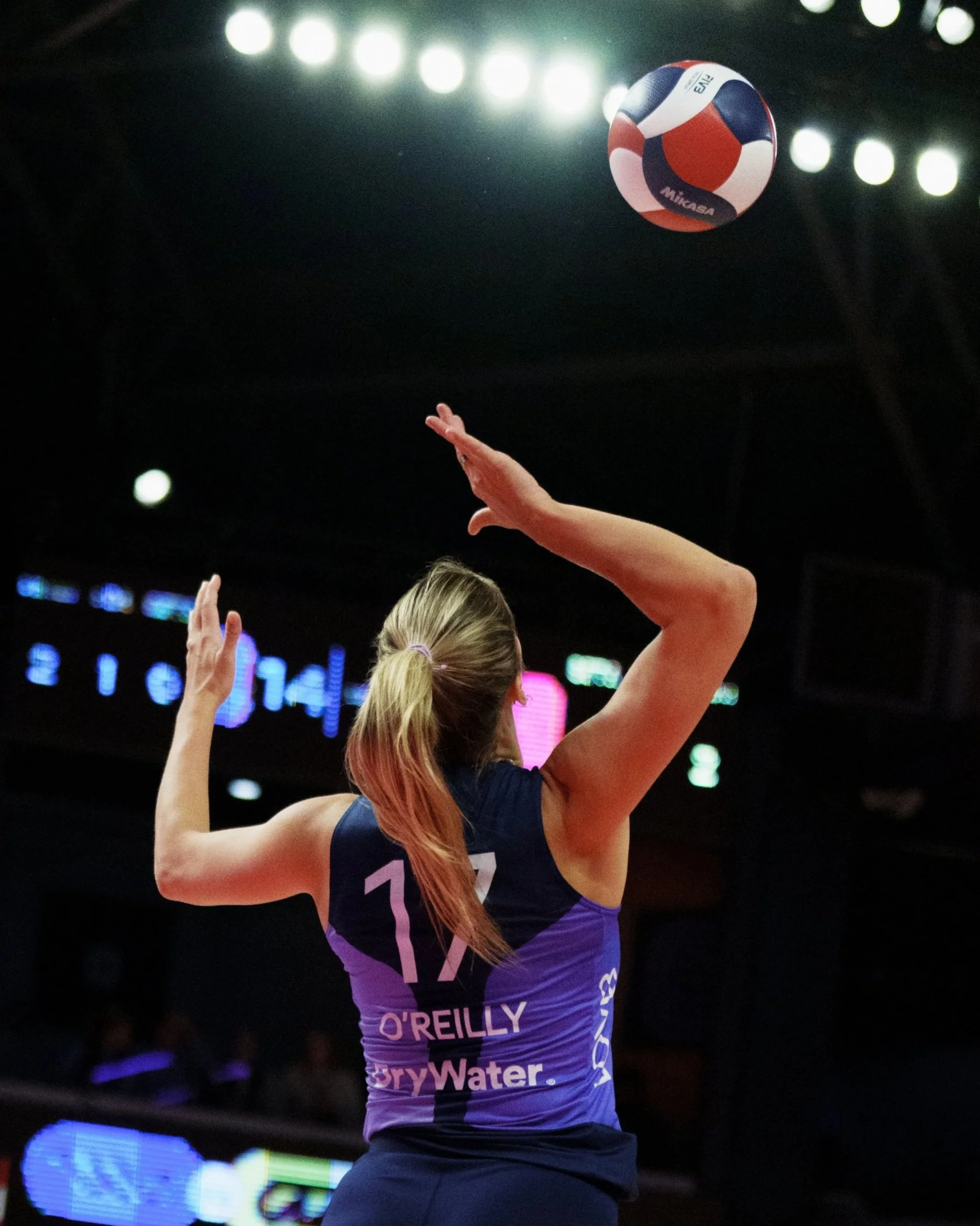 Female volleyball player with blonde hair tied in a ponytail, wearing a purple jersey with the number 17, preparing to serve in an indoor match. The ball is mid-air above her hand, and stadium lights are visible overhead.