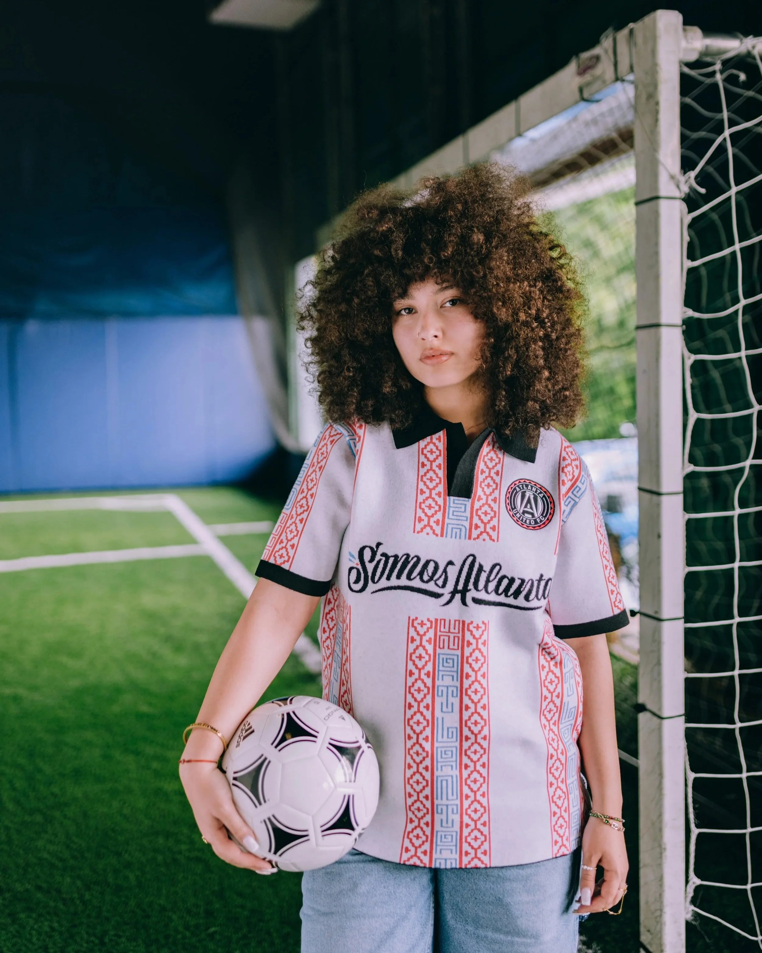 A young woman with curly hair wearing a sports jersey holding a soccer ball on a soccer field.