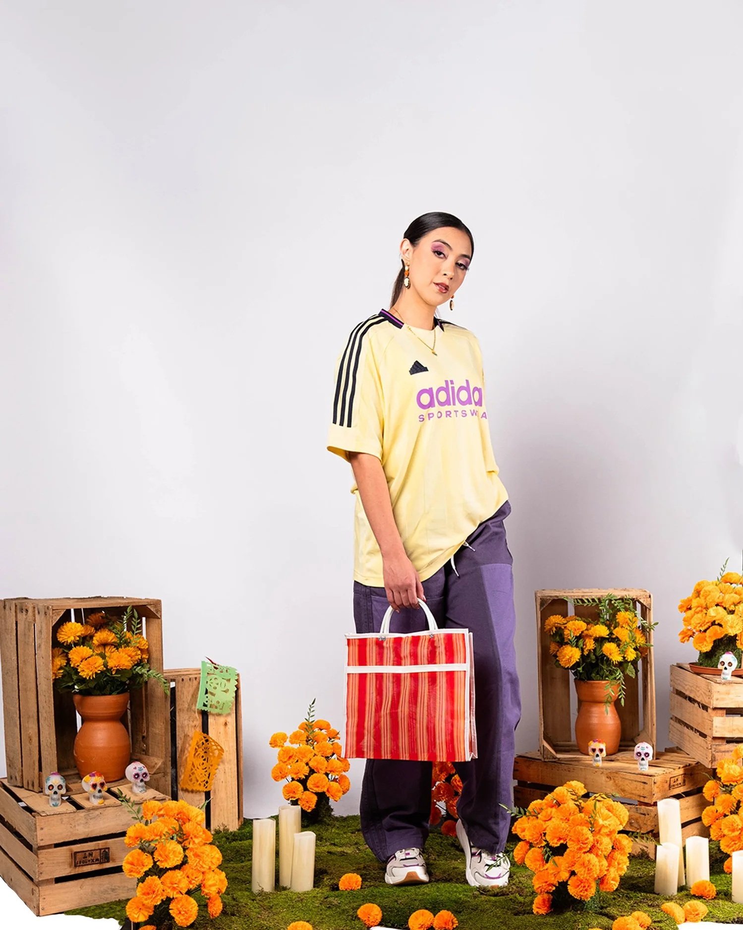 A woman standing in front of a backdrop decorated with orange marigolds, candles, and skull-themed ornaments. She is wearing a yellow Adidas jersey, purple pants, and white sneakers, holding a red and white striped bag. There are wooden crates filled with marigolds and skull decorations on either side of her.