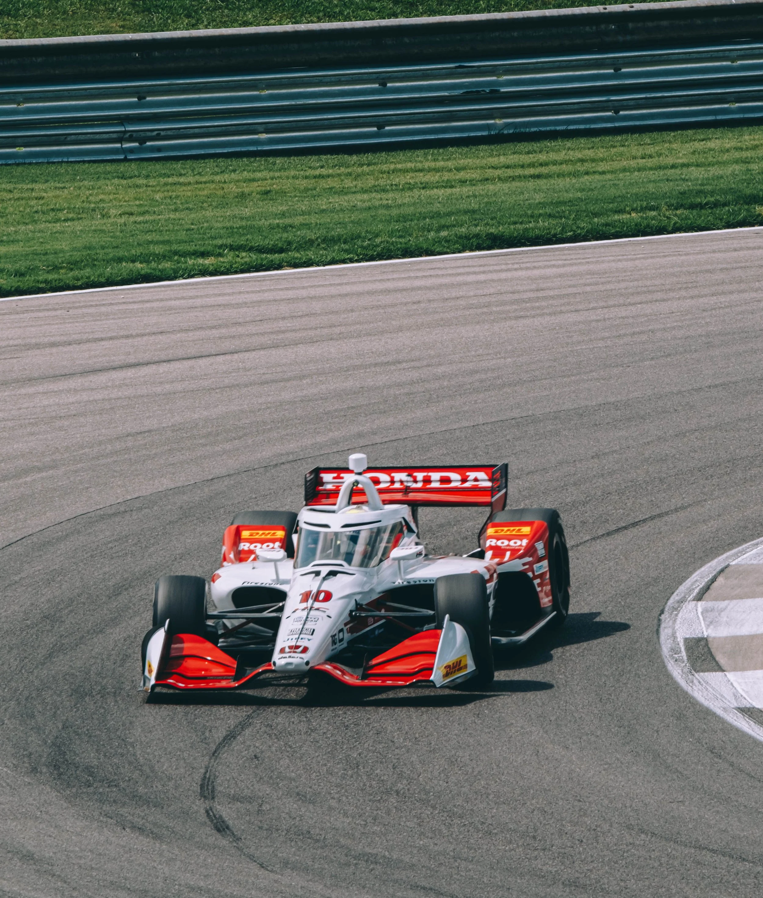 A race car on a race track. The car has Honda branding and sponsors like DHL and Root. It is navigating a curve on the track with tire marks visible behind it.