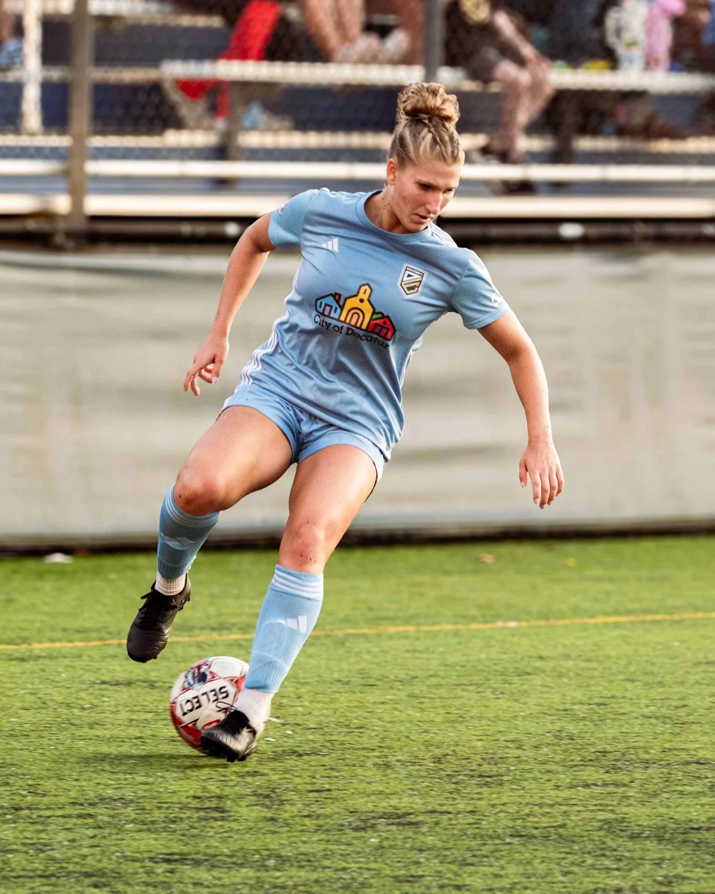 A female soccer player in a light blue uniform is on the field, kicking a white soccer ball with red and black details. She has her hair tied up in a bun and appears focused on the game. In the background, there are blurred spectators sitting on bleachers.