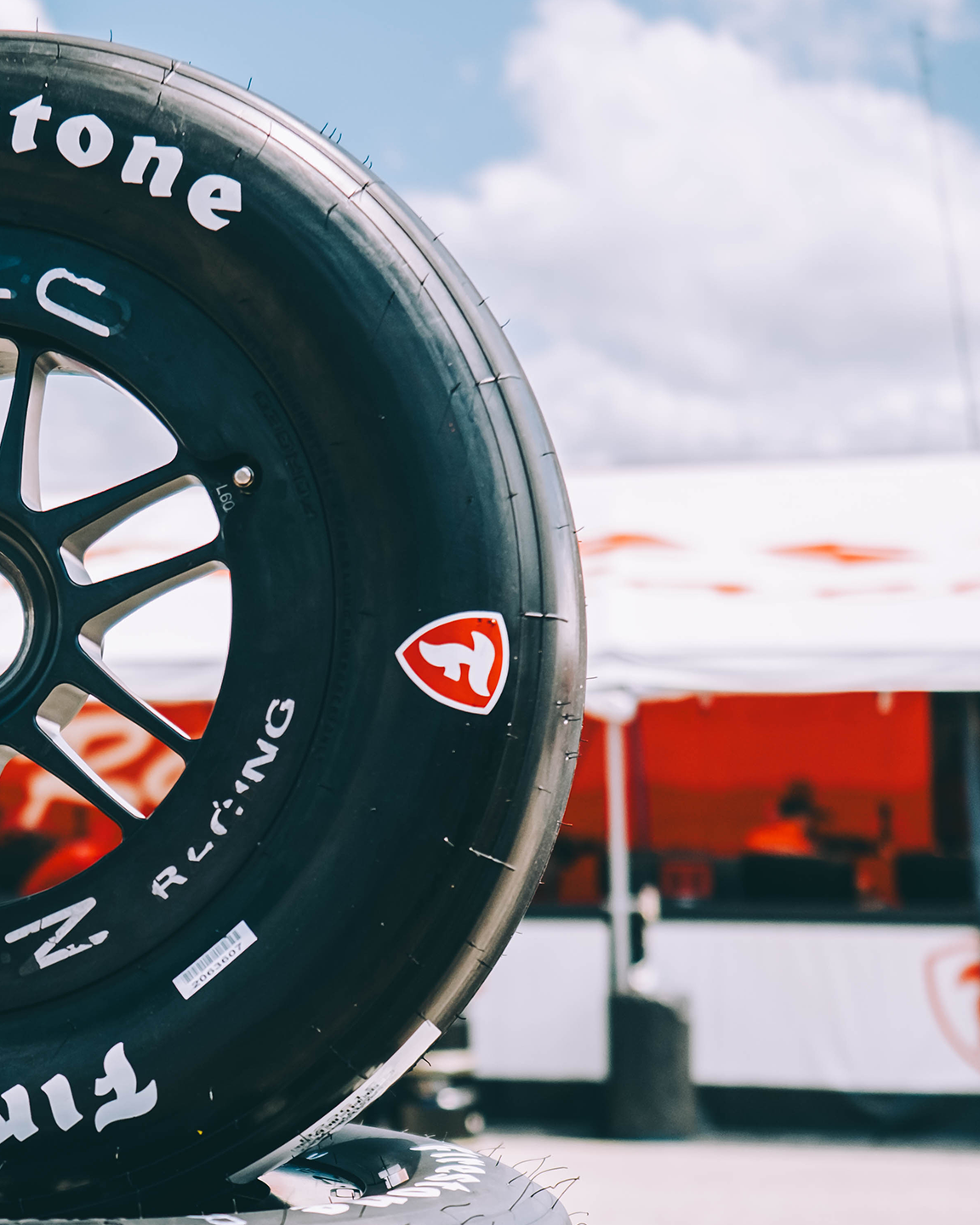 Close-up of a black racing tire with a red and white logo, set against a background of tents and a blue sky. firestone tires