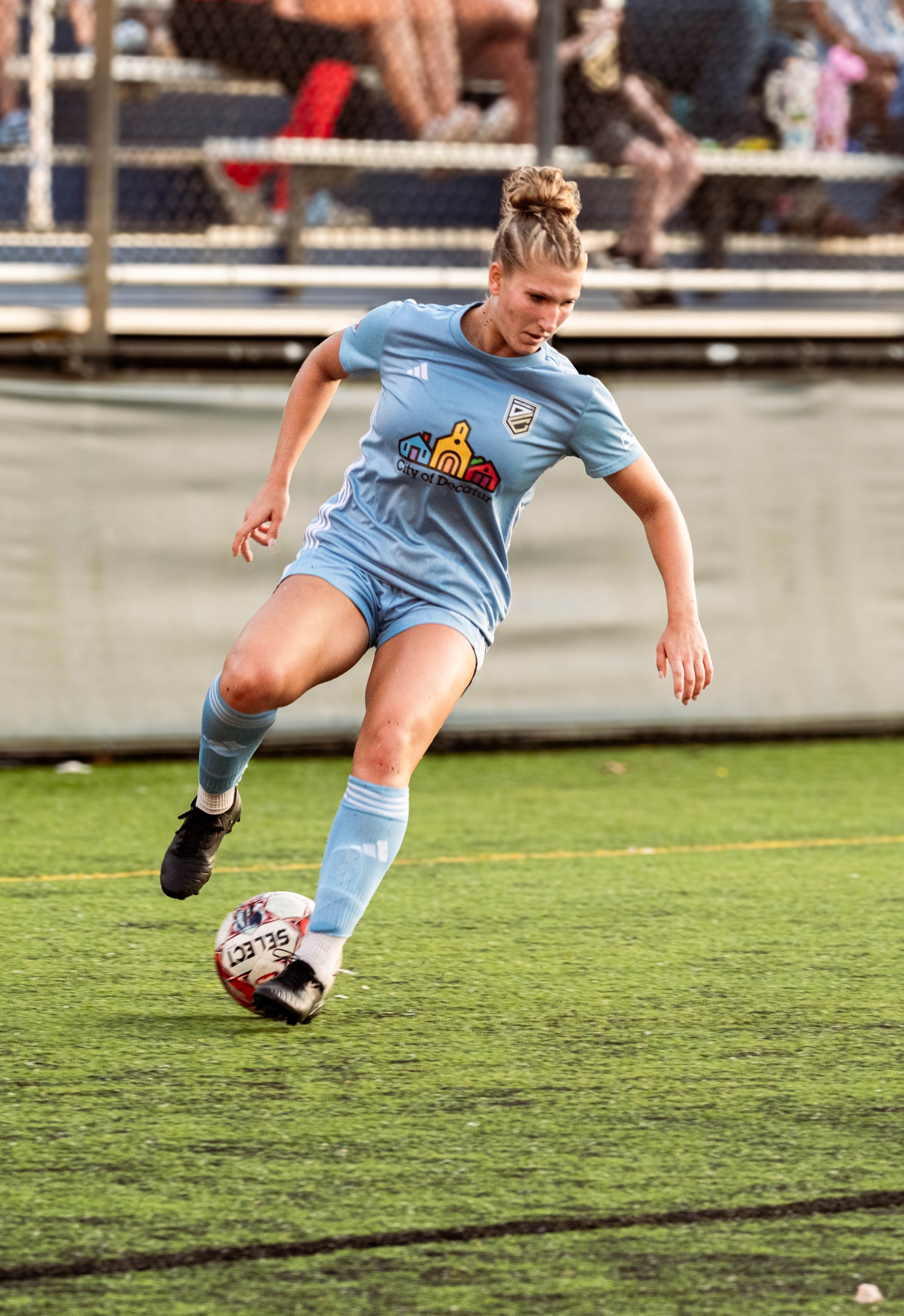 Female soccer player in a light blue uniform kicking a soccer ball on an outdoor field.