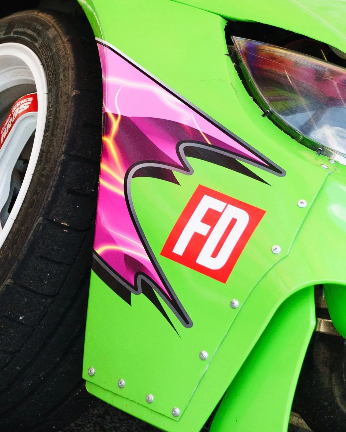 Close-up of the side of a race car with a bright green body, colorful graphics, and a large red and white sponsor logo, showing part of a tire and wheel.