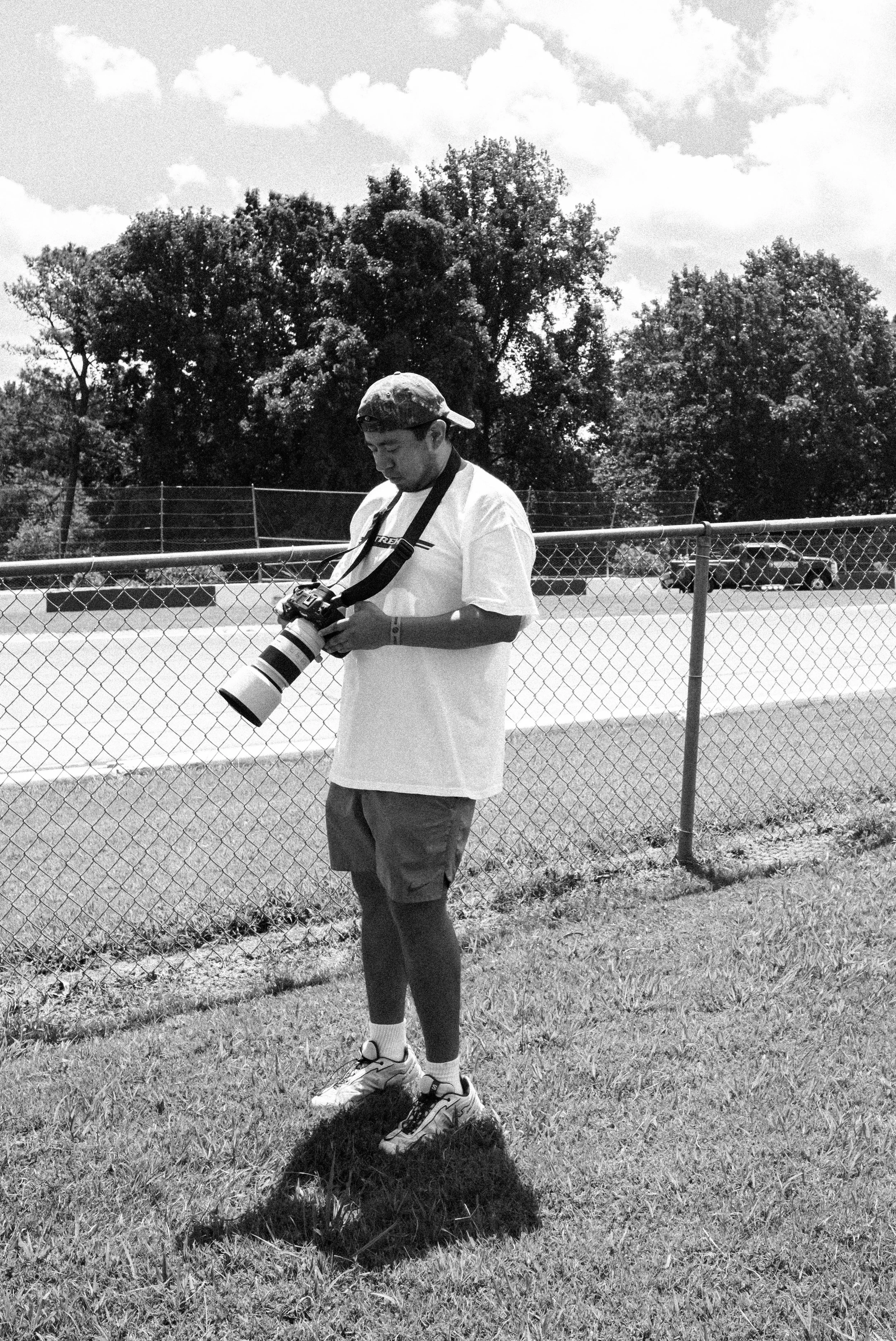 Young man wearing a cap, t-shirt, and shorts standing outdoors on grass, looking at a camera with a long lens, next to a chain-link fence, with trees and parked cars in the background, under a partly cloudy sky.