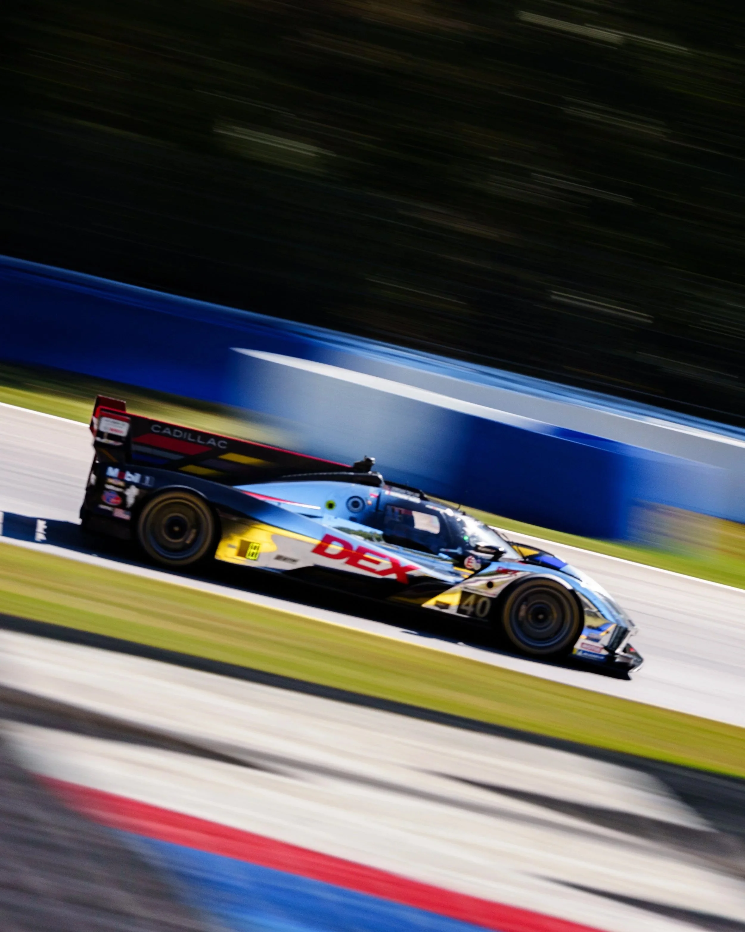 A race car on a racetrack at night, moving at high speed with motion blur. The car has decals and sponsorship logos, including 'DEX' and 'Cadillac'.