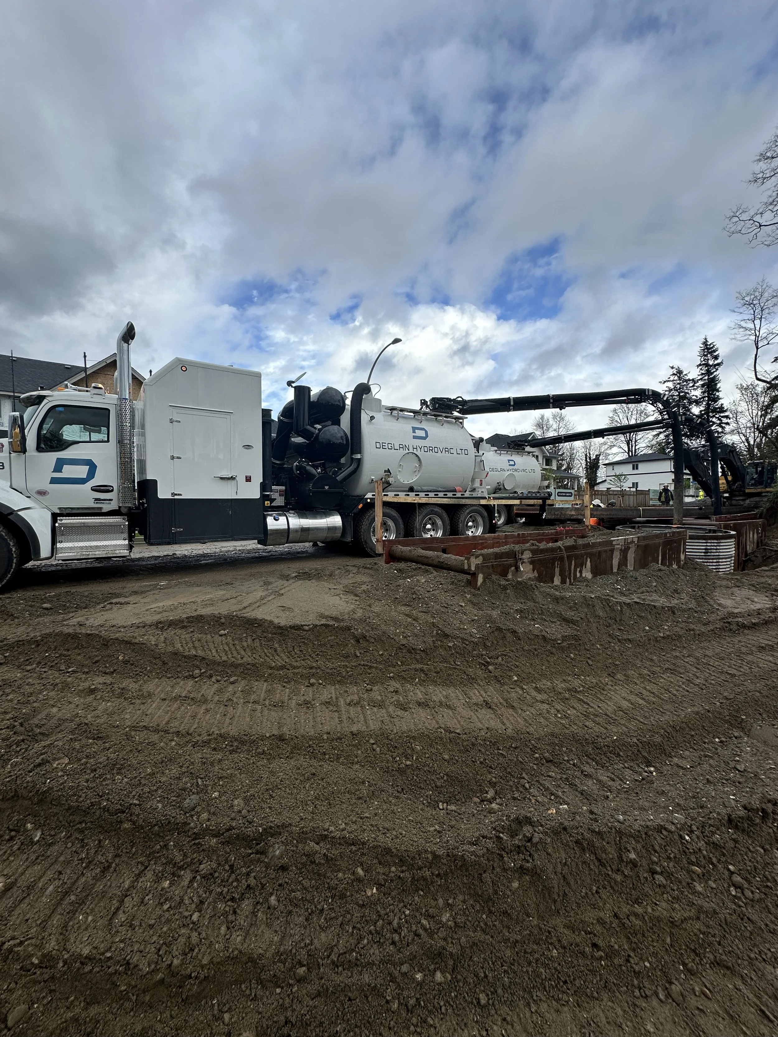 Construction site with a large white vacuum truck labeled Deldan Hydrovac Ltd. parked on dirt ground under a cloudy sky.