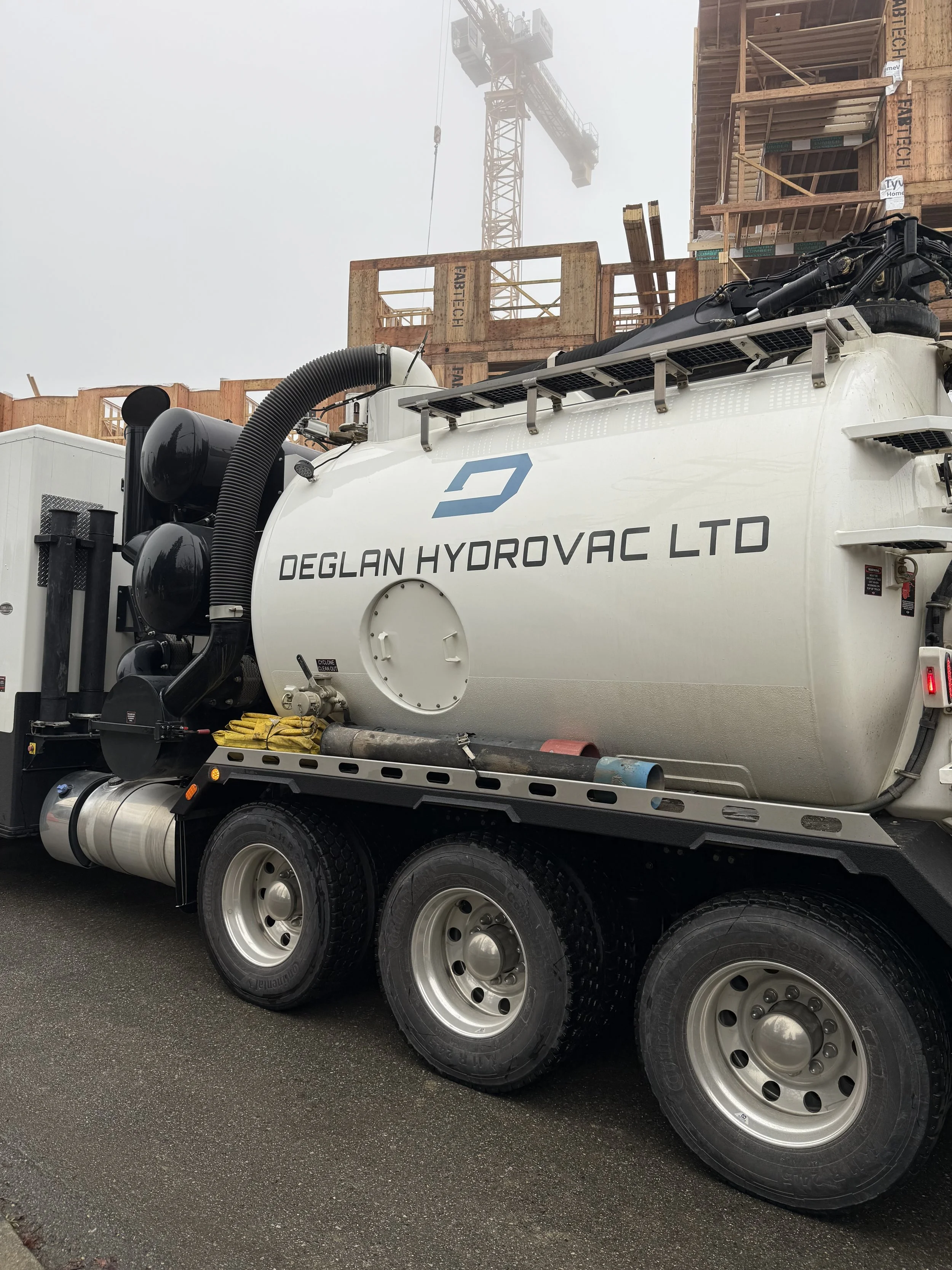 A large white vacuum truck with the label 'Deglan Hydrovac Ltd' parked in front of a construction site with a crane and unfinished building.