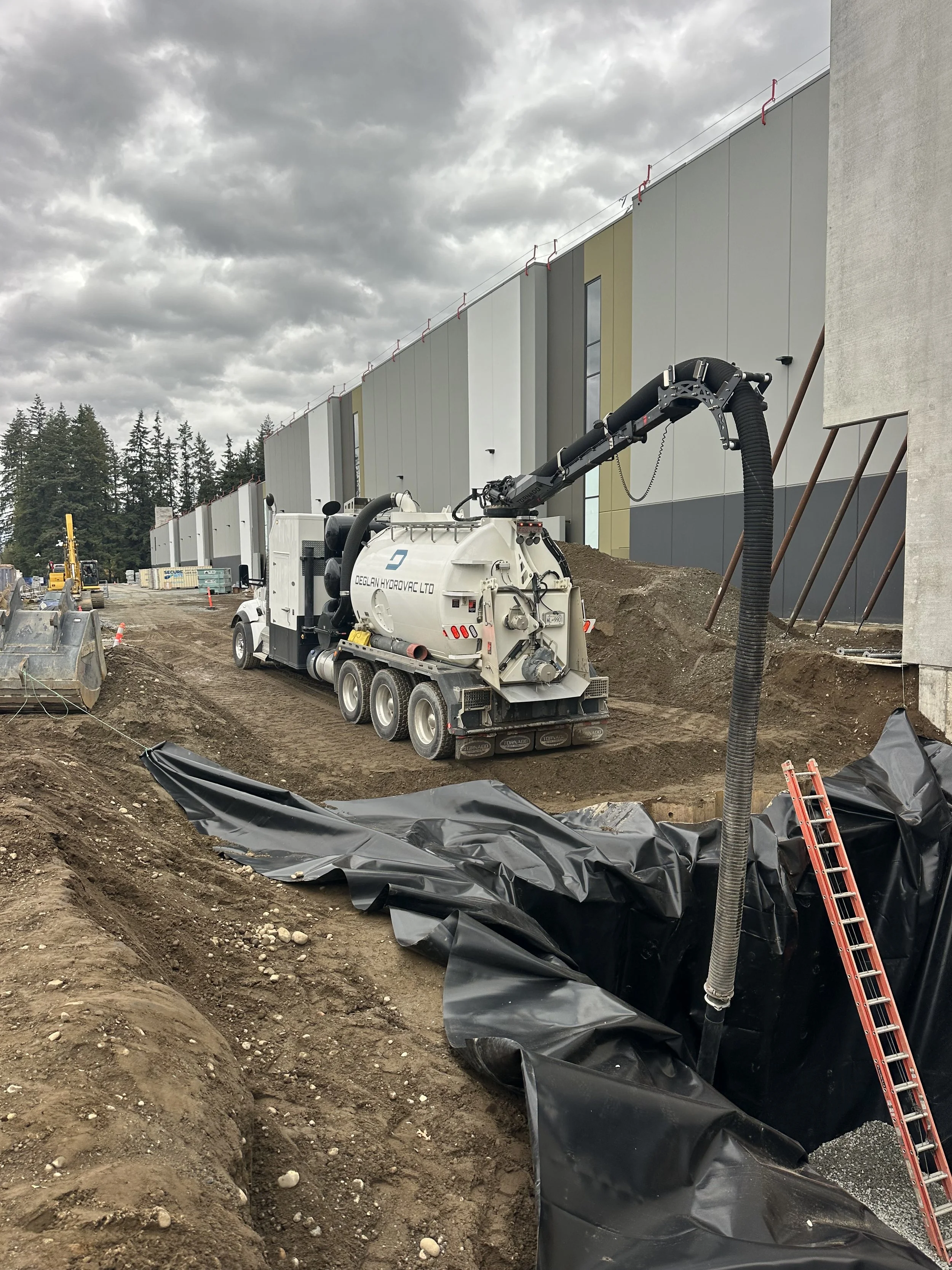Construction site with a vacuum truck spraying a black liquid onto dirt, a ladder leaning into a large excavated area, and a large modern building in the background under cloudy skies.