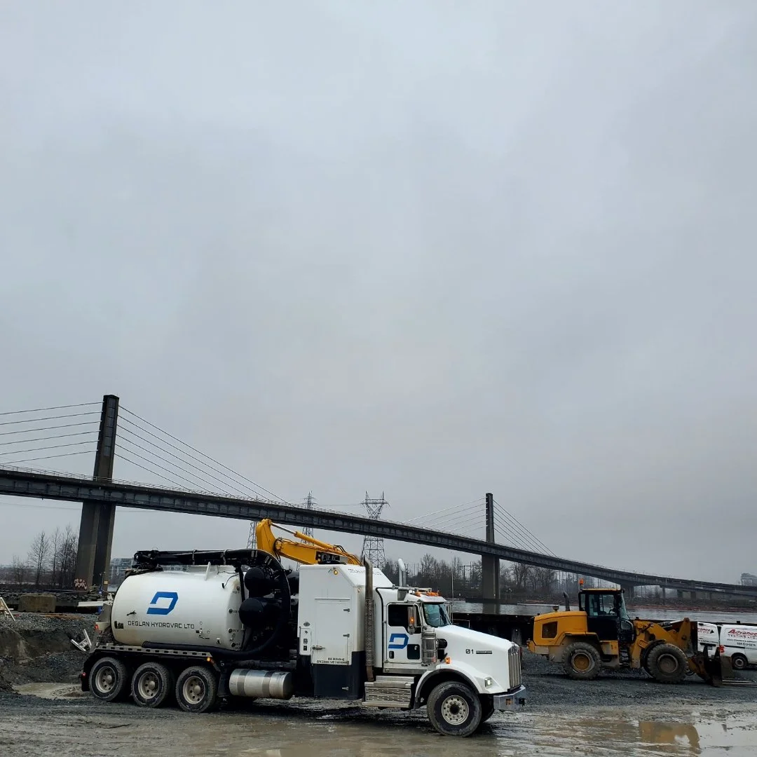 Construction site with a large white vacuum truck and a yellow bulldozer under an overcast sky, with a bridge and power lines in the background.