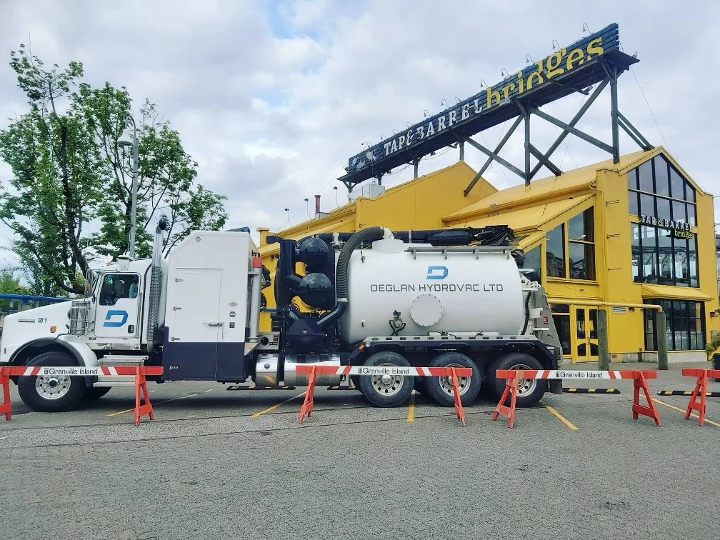 A white truck with a large vacuum tank, labeled 'DEGLAN HYDROVAC LTD,' parked in front of a yellow building with large glass windows and a sign that reads 'TAP & BRIDGES.' The area is cordoned off with orange and black barricades.
