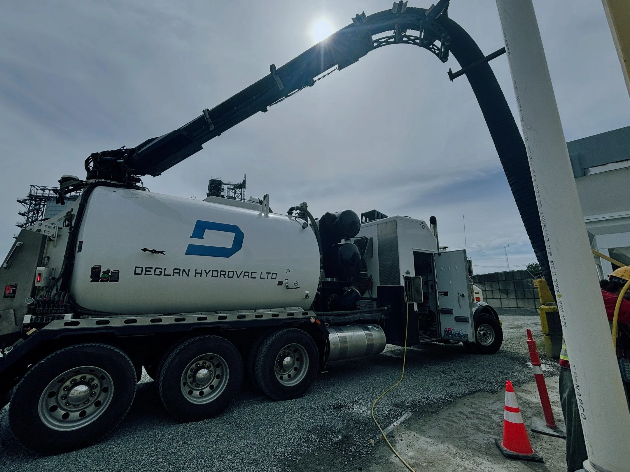 A concrete pump truck with the logo and name 'Deglan Hydrovac Ltd' on the tank, extending its long black boom with a hose, parked at a construction site near a gravel area under a cloudy sky.