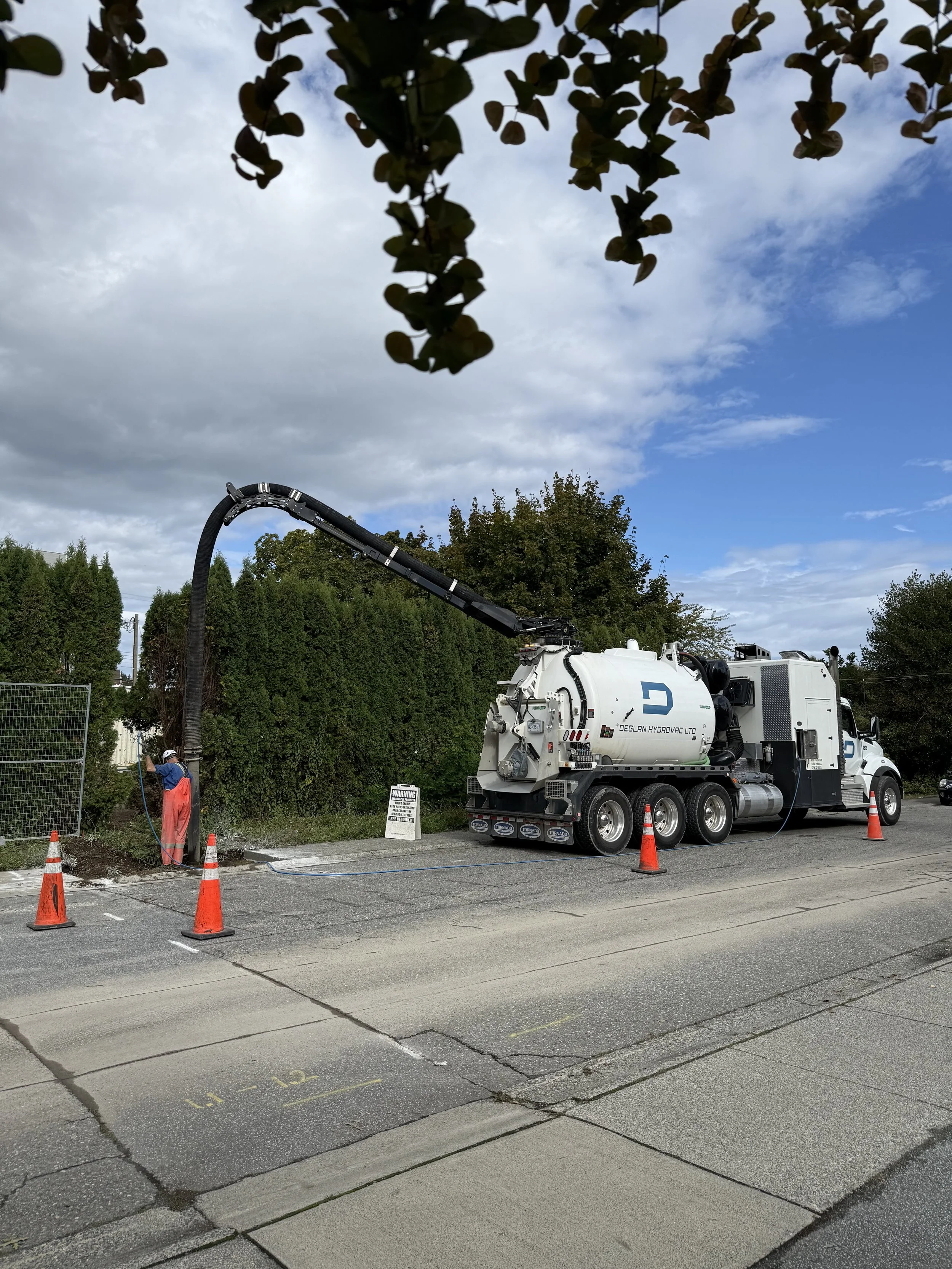 A worker in orange safety gear stands beside a street worker vacuum truck, with garbage excavation in progress, surrounded by four orange traffic cones on a city street with trees and cloudy sky in the background.